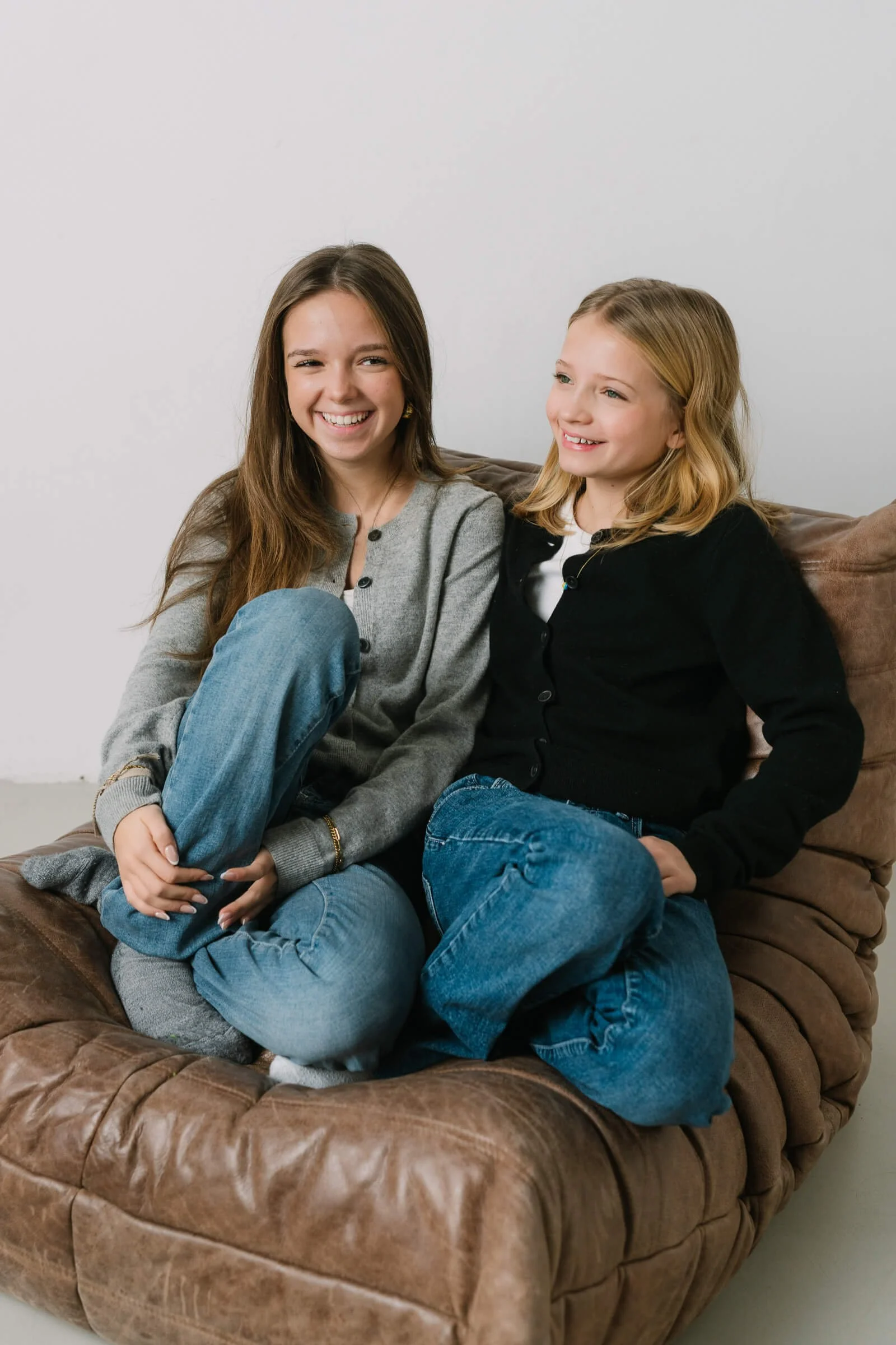 Two young girls sitting together on a brown leather couch, smiling and enjoying each other's company.