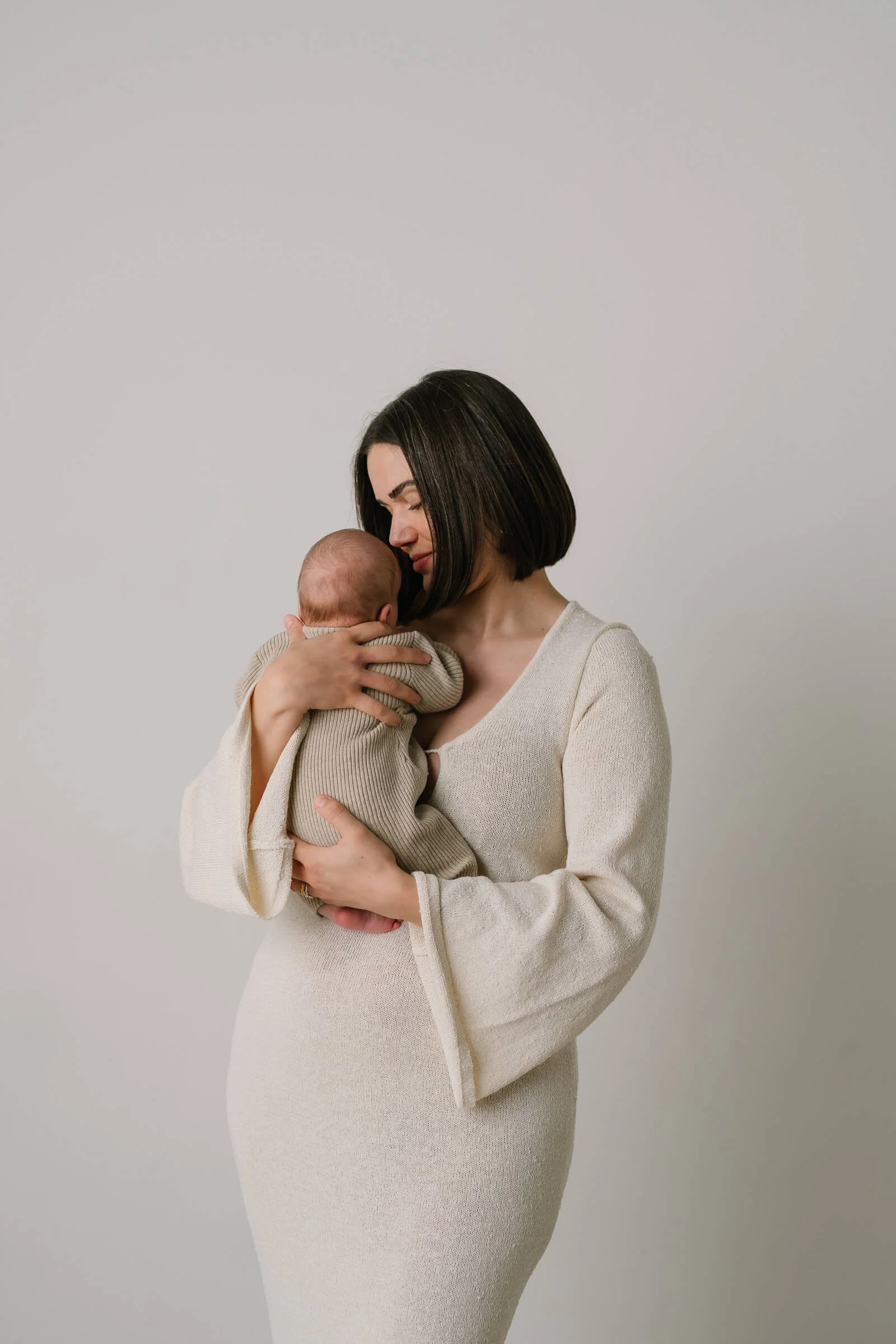 A woman holding a baby close to her face, sharing a tender moment indoors with a plain background.