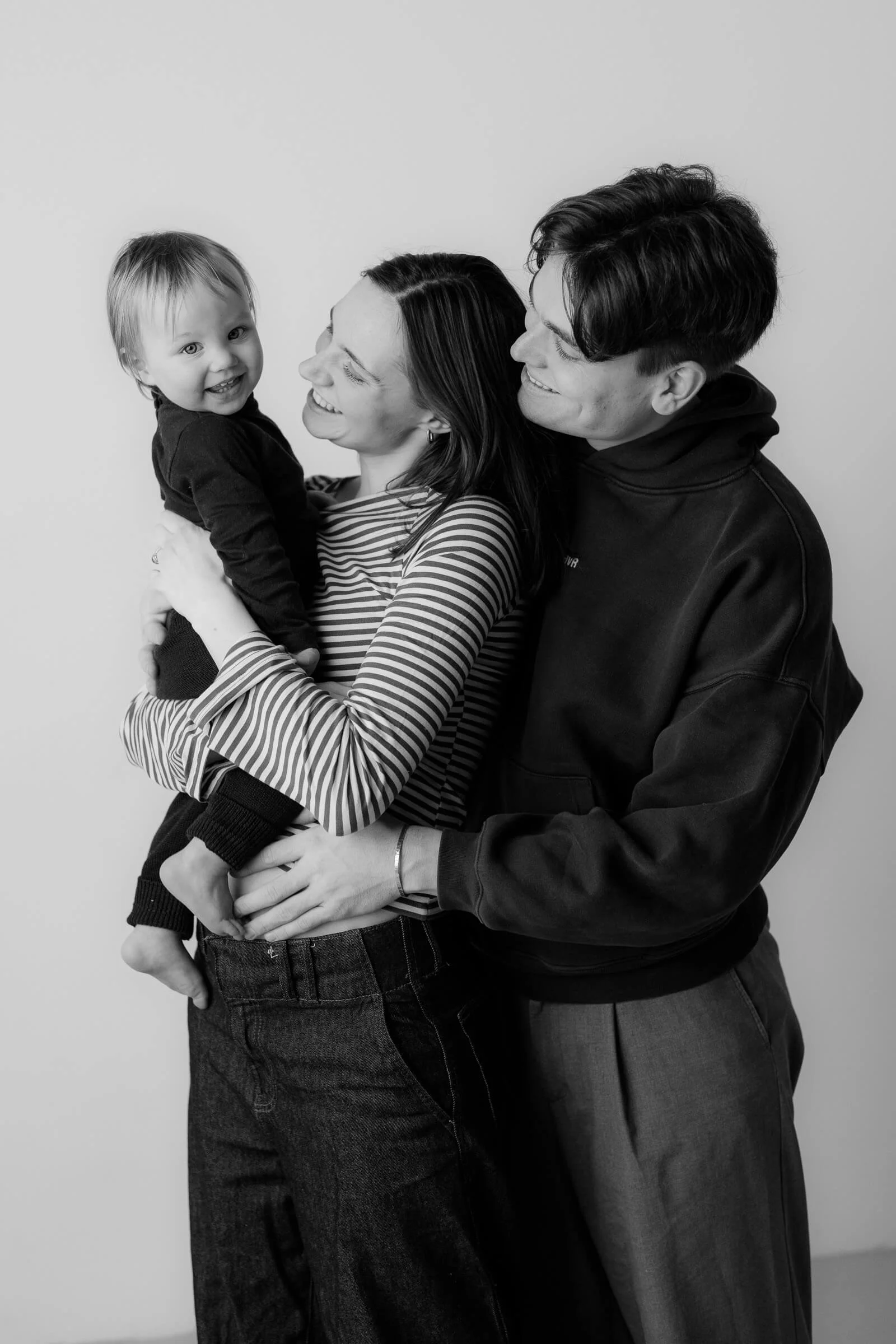 A black and white photo of a smiling family of three. The woman holds a young child, and the man embraces them both, all smiling at each other.