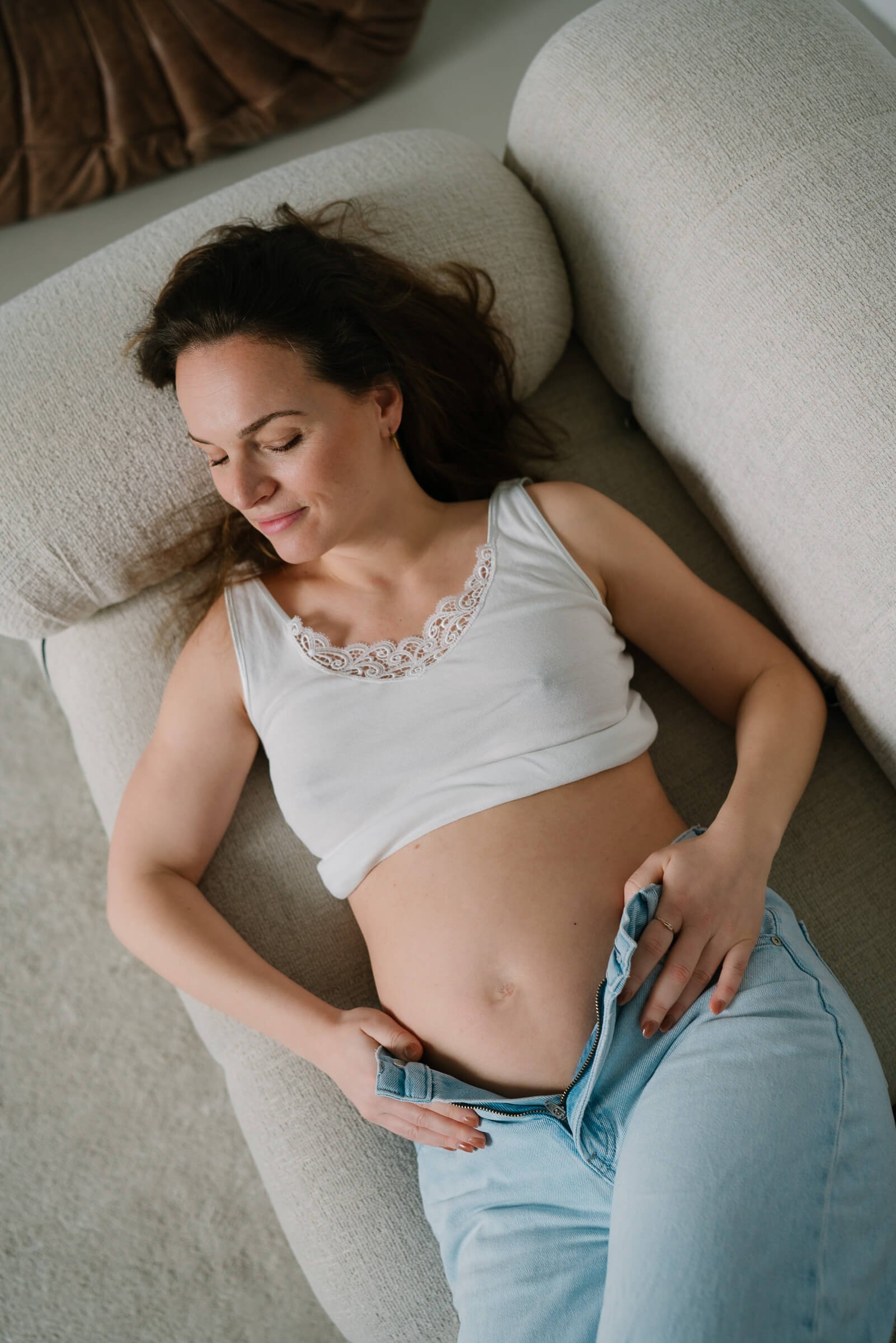 A woman with dark hair lying on a beige sofa with eyes closed, wearing a white tank top with lace trim and light blue jeans unzipped, resting her hand on her lower abdomen.