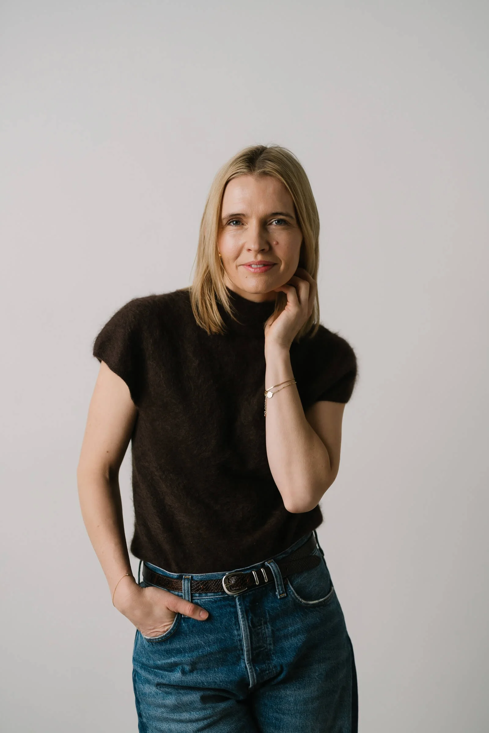 A woman with blonde hair wearing a black turtleneck sweater and blue jeans, standing against a plain gray background, looking at the camera with a slight smile.