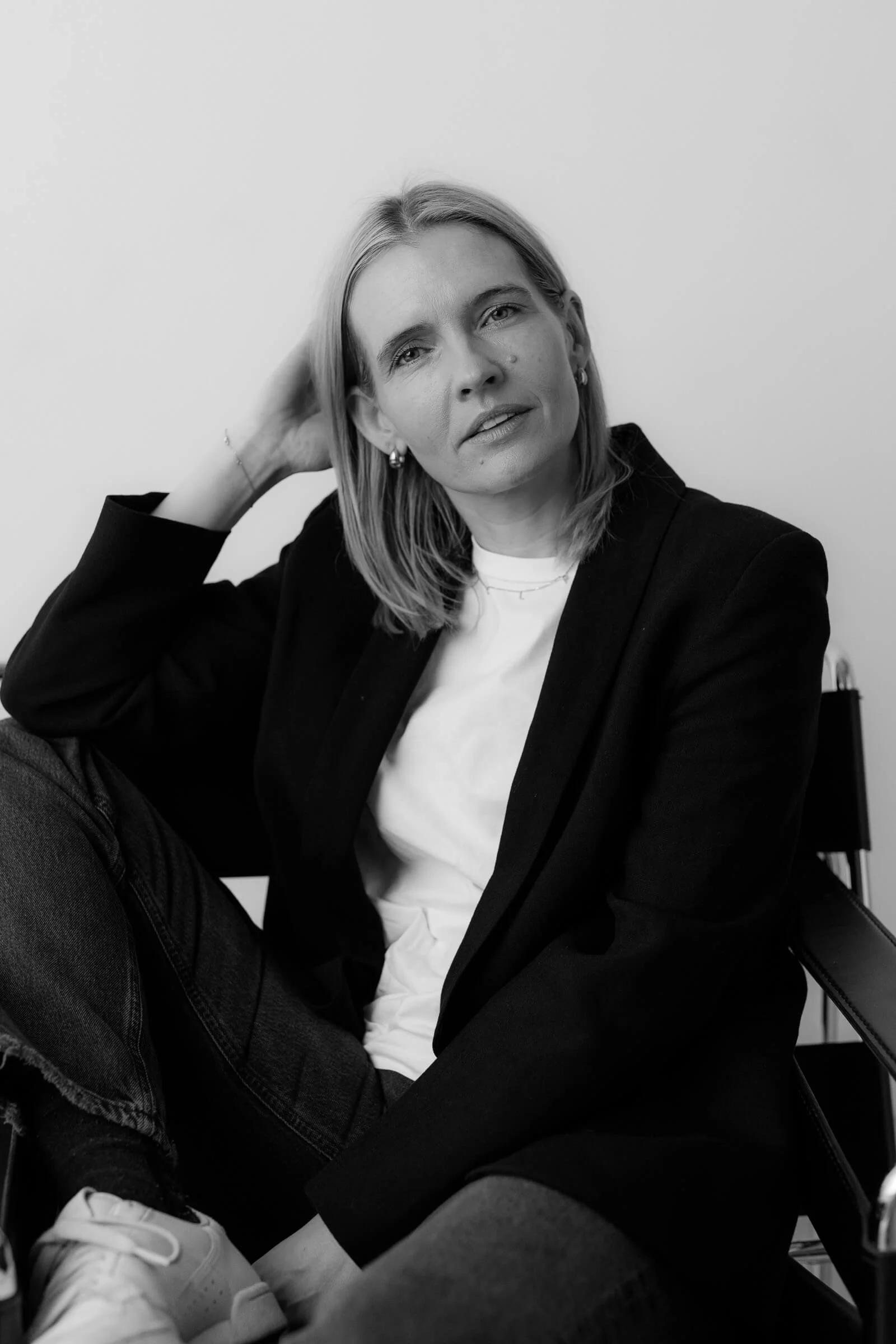 A black-and-white photo of a woman with shoulder-length blonde hair, wearing a black blazer, white t-shirt, and dark jeans, sitting on a chair against a plain light-colored wall, with her left hand behind her head and looking at the camera.