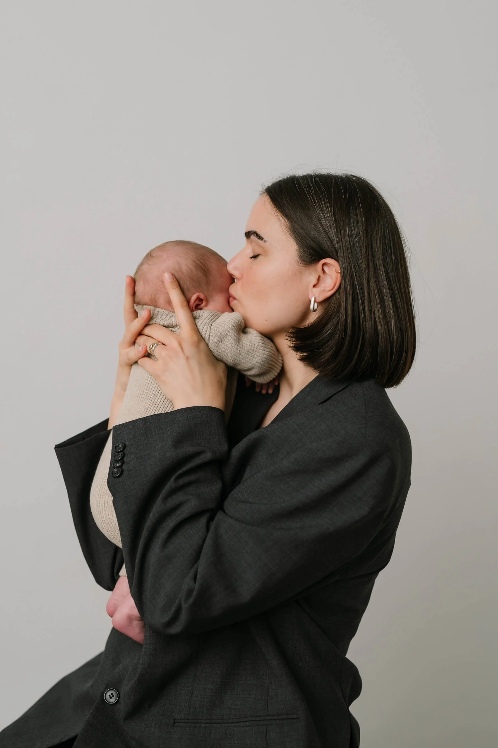 A woman in a black blazer holds a baby close, giving a gentle kiss on the baby's head, against a plain light gray background.