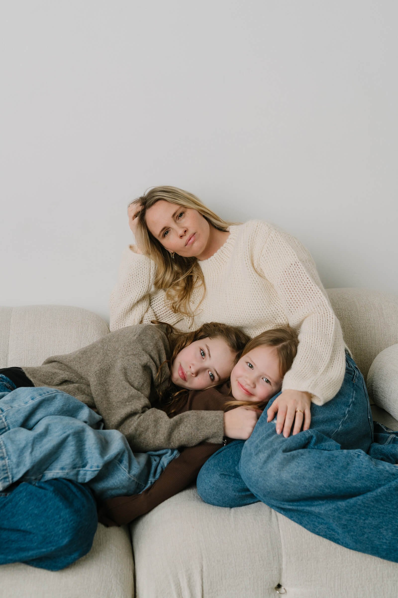 A woman sitting on a beige couch with two children, all in casual clothing, against a plain white wall. The woman has blonde hair and is wearing a cream knit sweater. The children, one with brown hair and the other with red hair, are lying on the wom