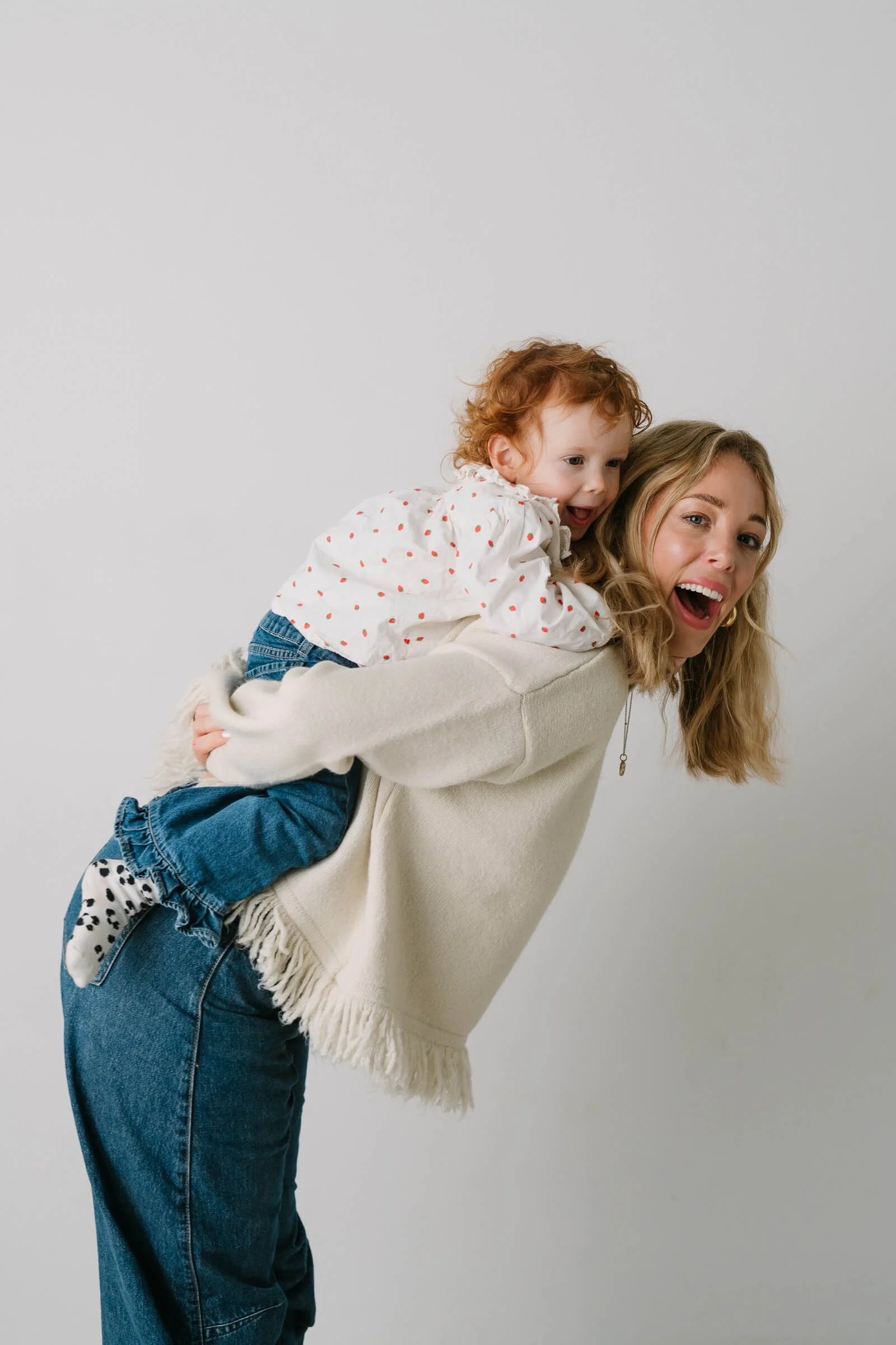 A woman with blonde hair and a white sweater carrying a young girl with red curly hair on her back, both smiling and looking surprised, against a plain white background.