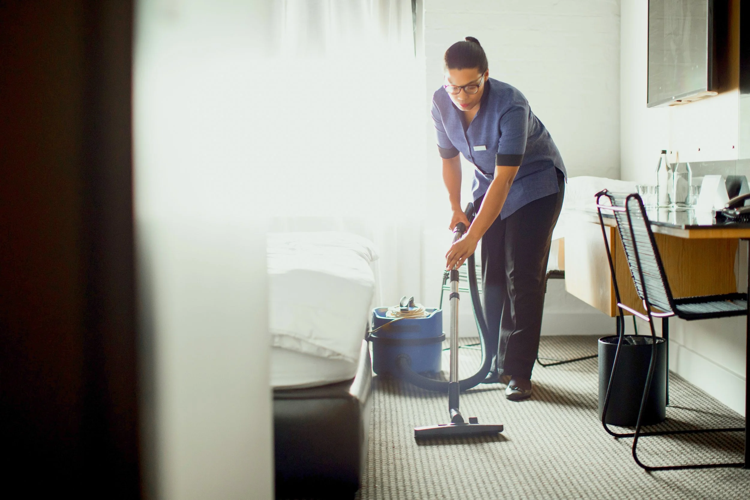 A woman wearing glasses and a gray uniform is vacuuming a hotel room, with chairs and a desk in the background.