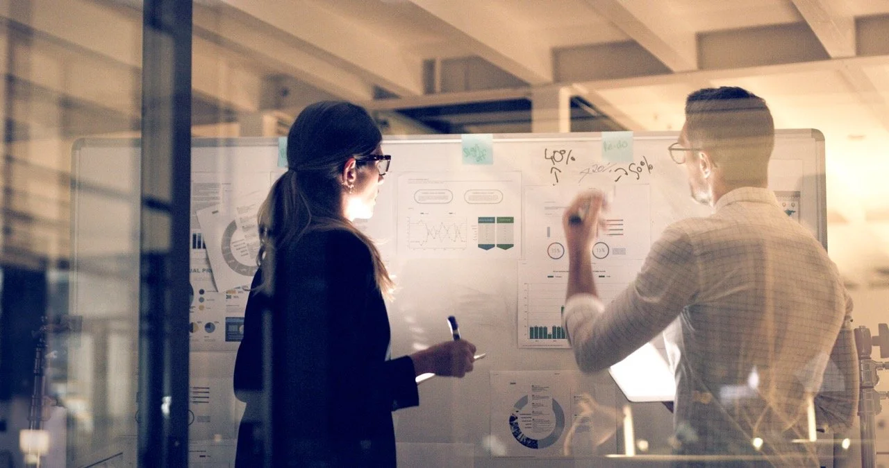 Two professionals, a woman and a man, discussing data displayed on a whiteboard filled with charts and graphs in an office.