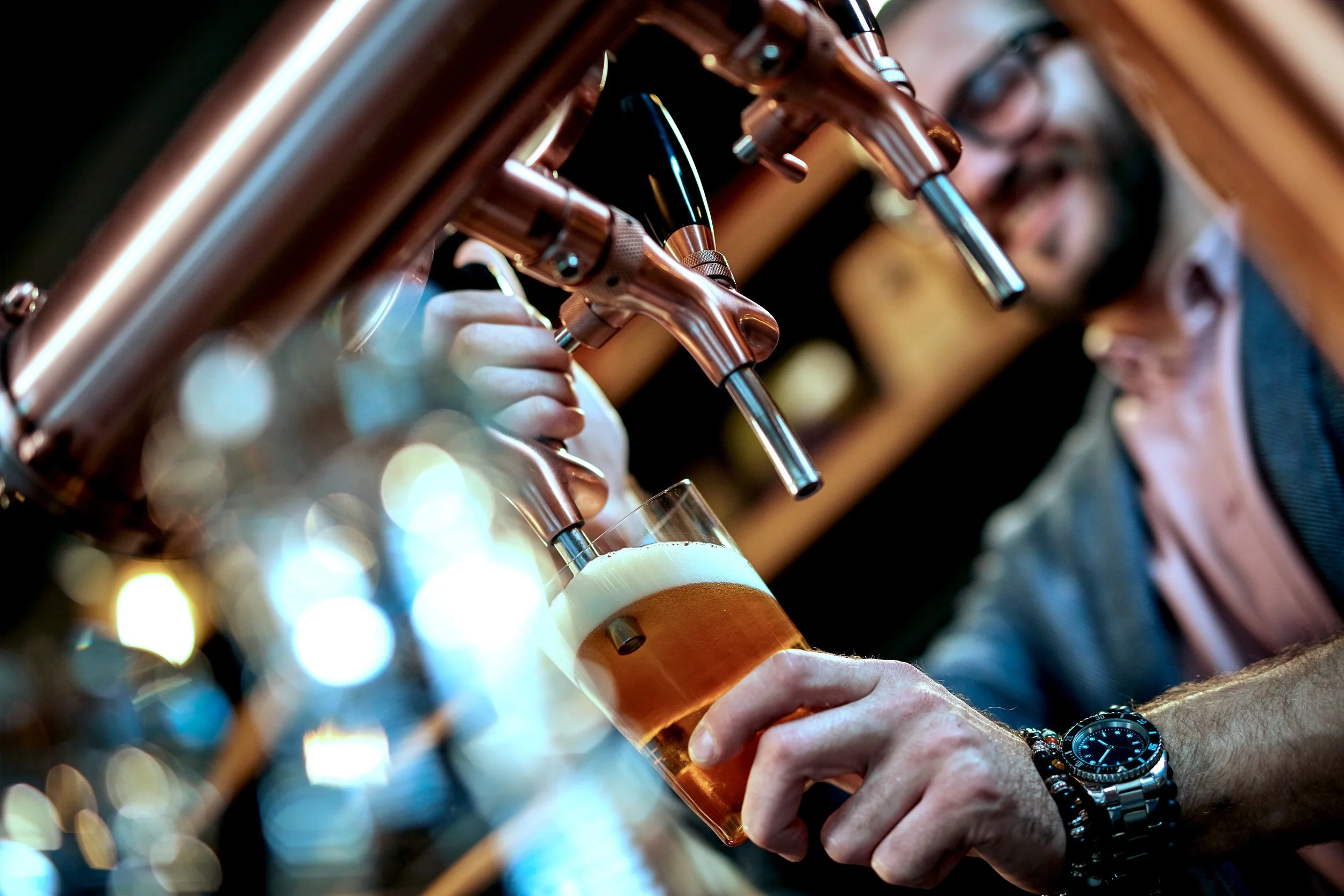 Close-up of a person pouring a beer from a tap faucet into a glass, with a woman in glasses blurred in the background.