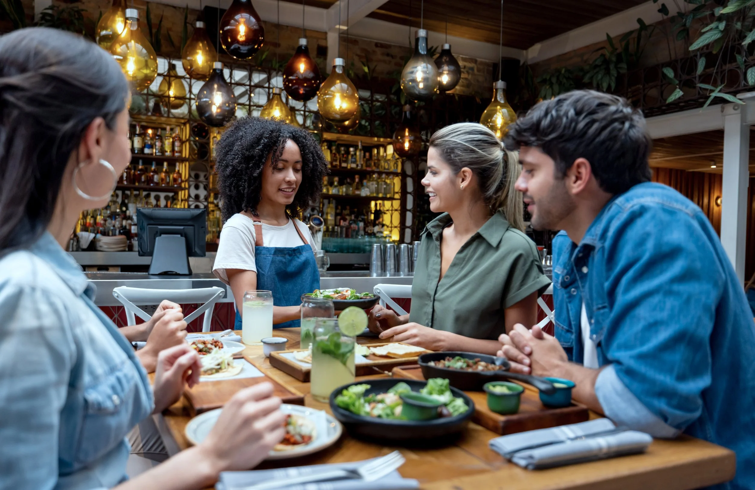 A group of five diverse young adults sitting at a wooden table in a restaurant, enjoying a meal together. There are various dishes, salads, and drinks on the table, with a bar and decorative hanging lights in the background.