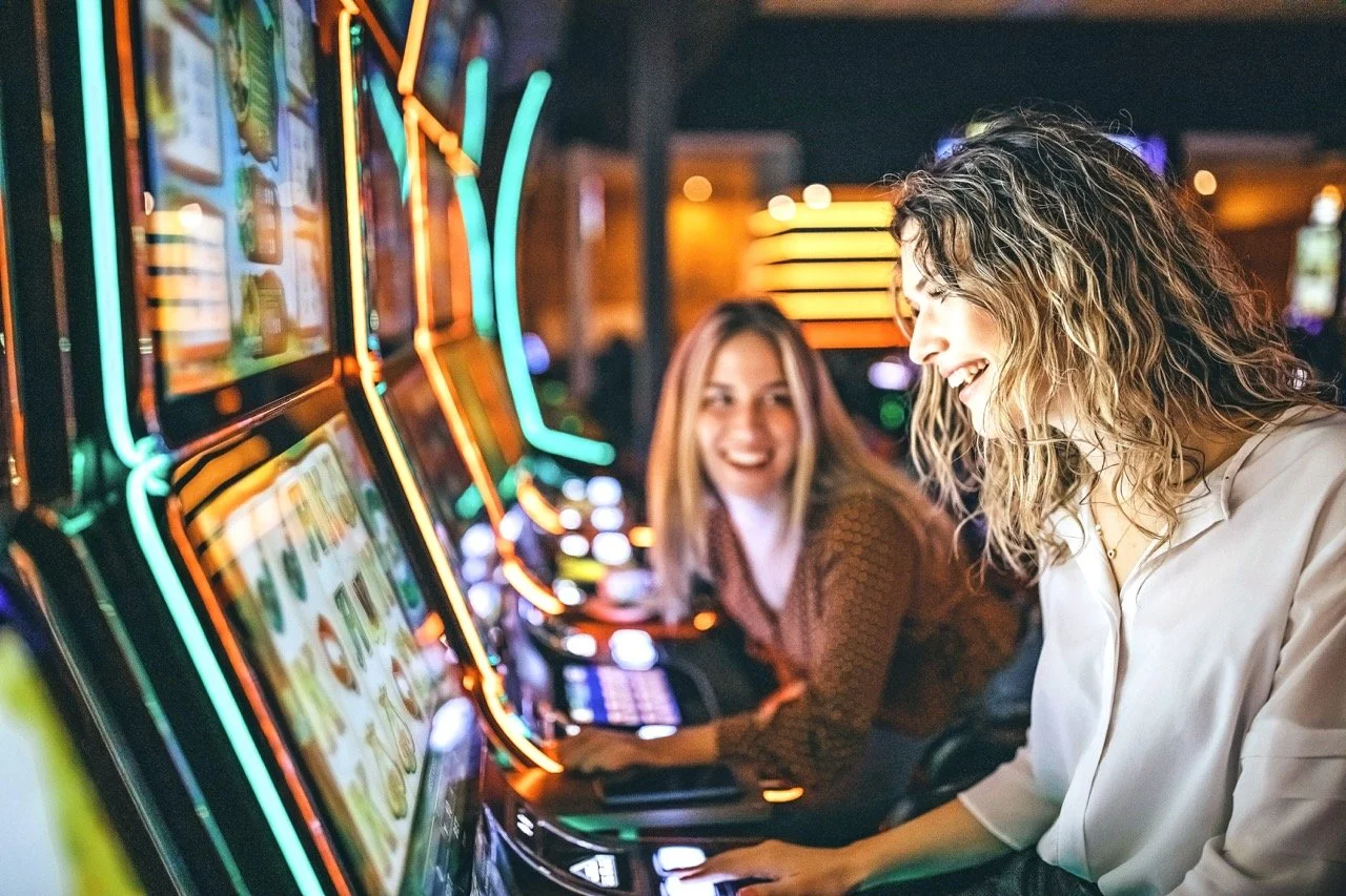 Two women playing slot machines at a casino at night, smiling and enjoying the game.