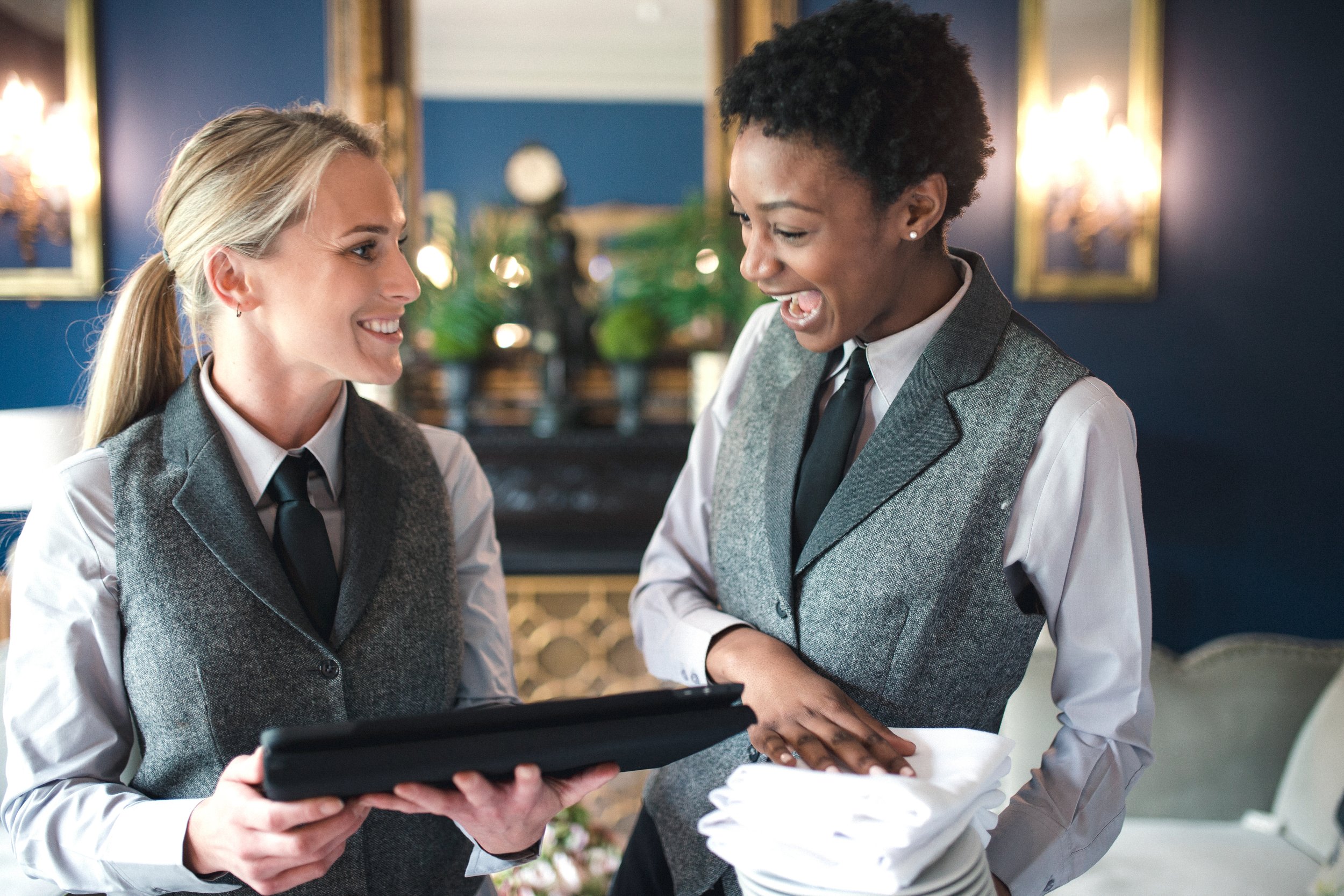 Two waitresses dressed in gray vests, white shirts, and black ties shaking hands and chatting in a restaurant with blue walls and gold-framed mirrors.