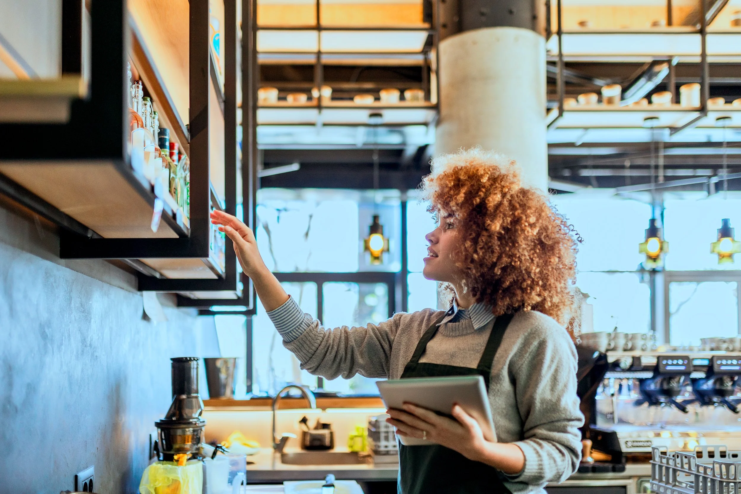 A woman with curly hair wearing a striped shirt and apron reaching for an item on a shelf in a modern café or coffee shop.