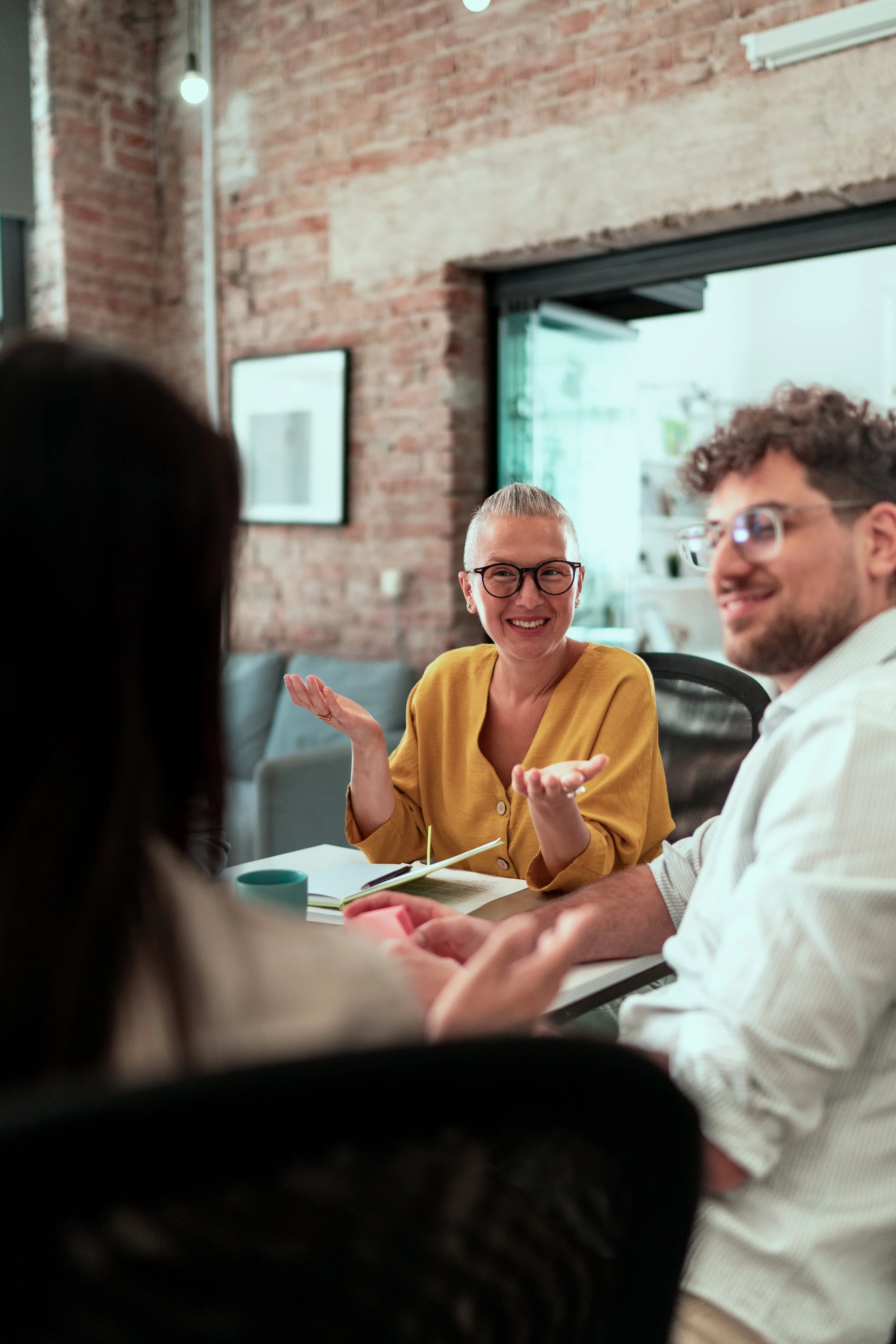 A group of people having a discussion in a modern, industrial-style office with exposed brick walls and large windows.