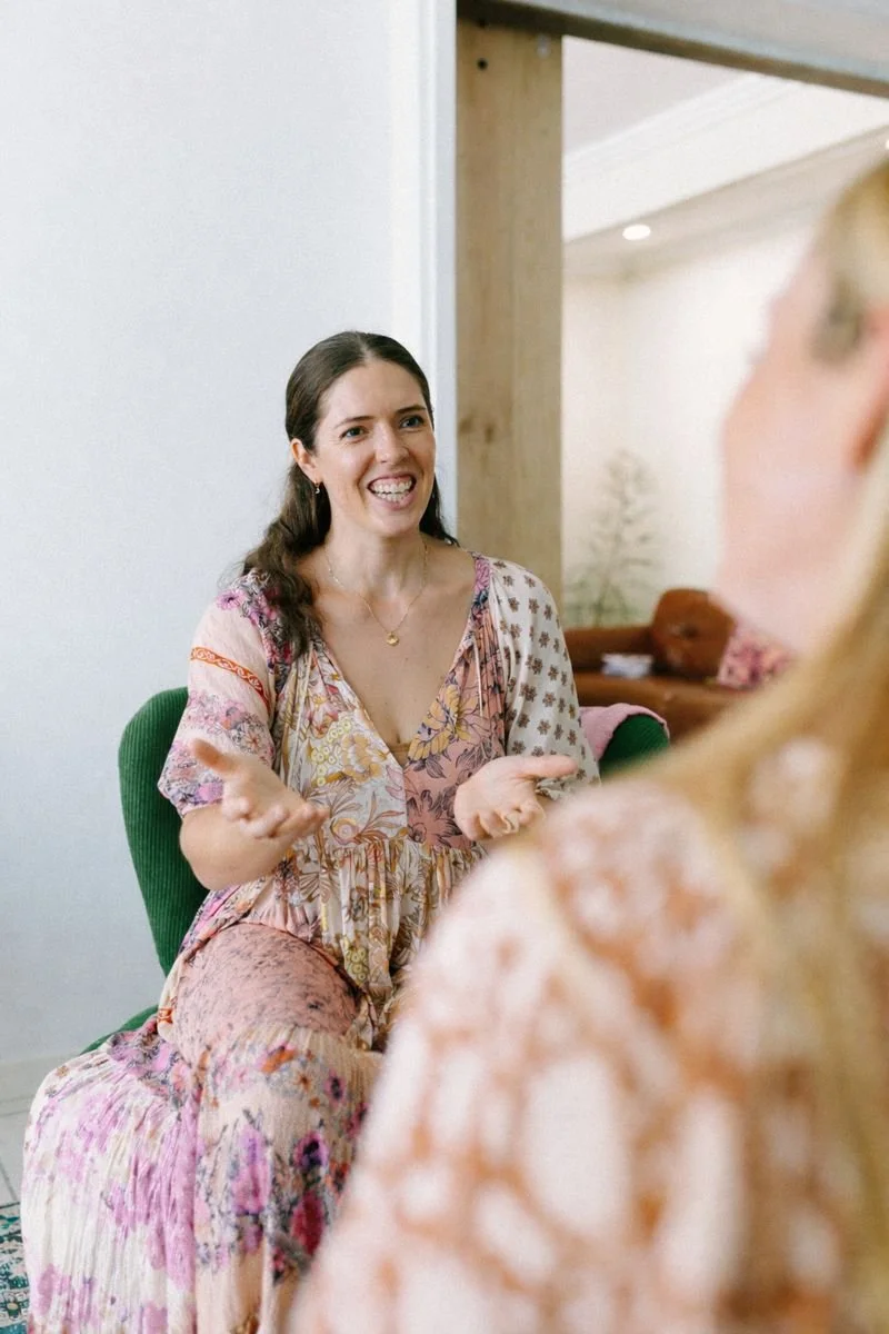 A woman with long dark hair, wearing a floral dress, is sitting on a green chair, gesturing with her hands, and smiling while talking to another person in a cozy room.