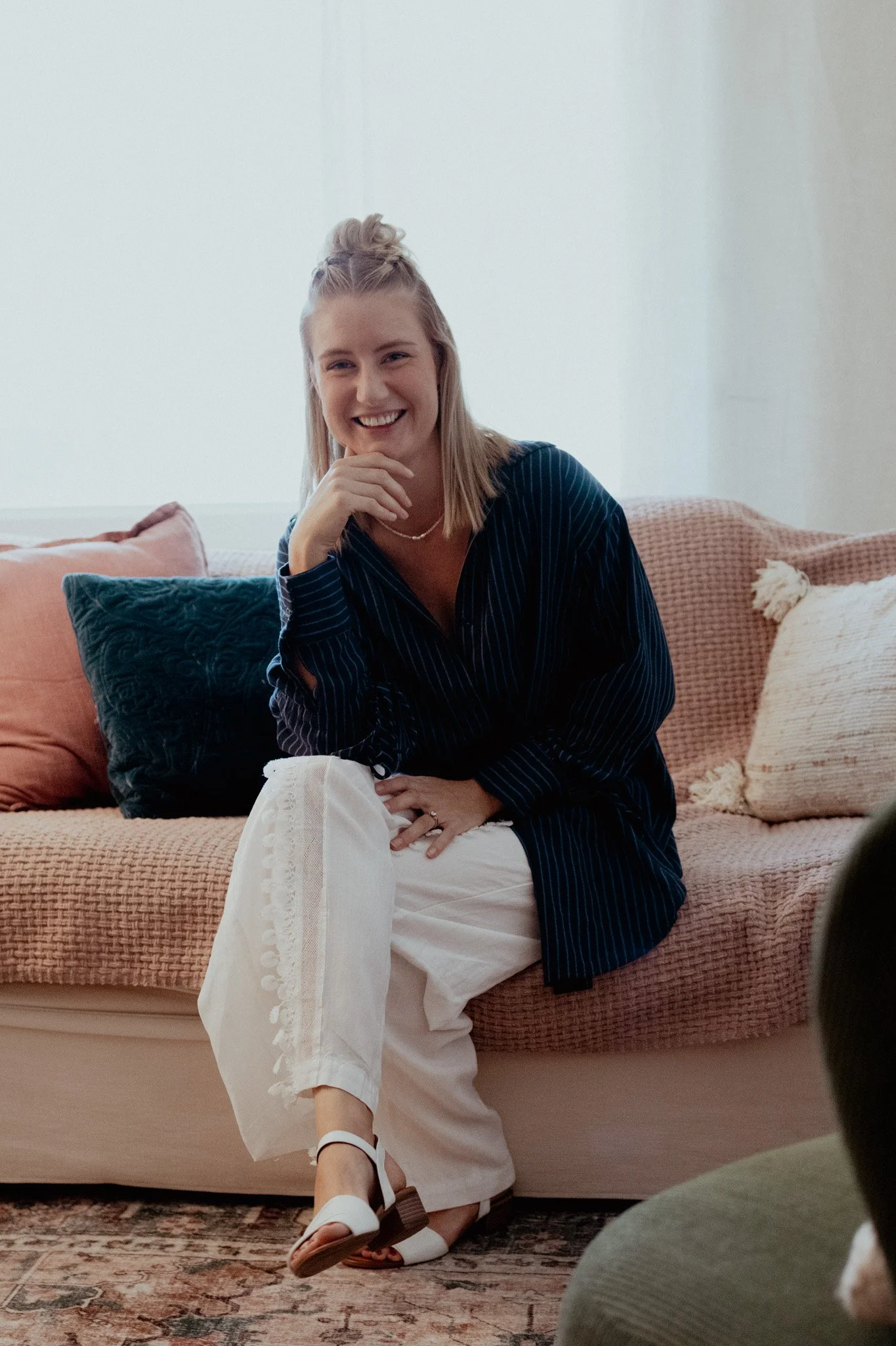 A woman sitting on a pink sofa, smiling, wearing a dark striped shirt and white pants.