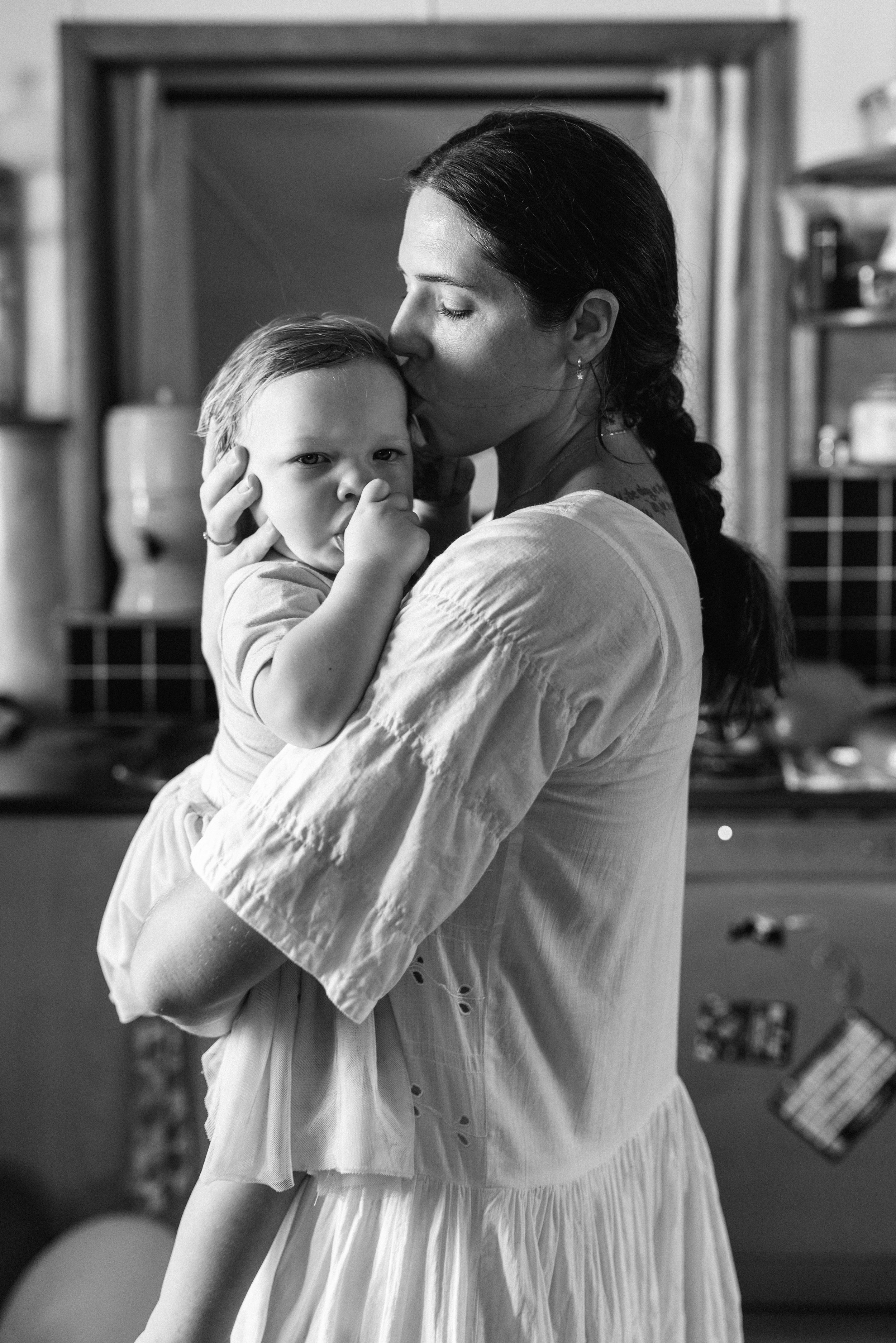 A woman holding a young child in her arms, with the woman kissing the child's forehead. The child has a curious expression and is touching their nose with their fingers. The woman has long, dark hair tied back and is wearing a white dress, while the child has short hair and a light-colored shirt. The background shows a kitchen setting with shelves and various household items.