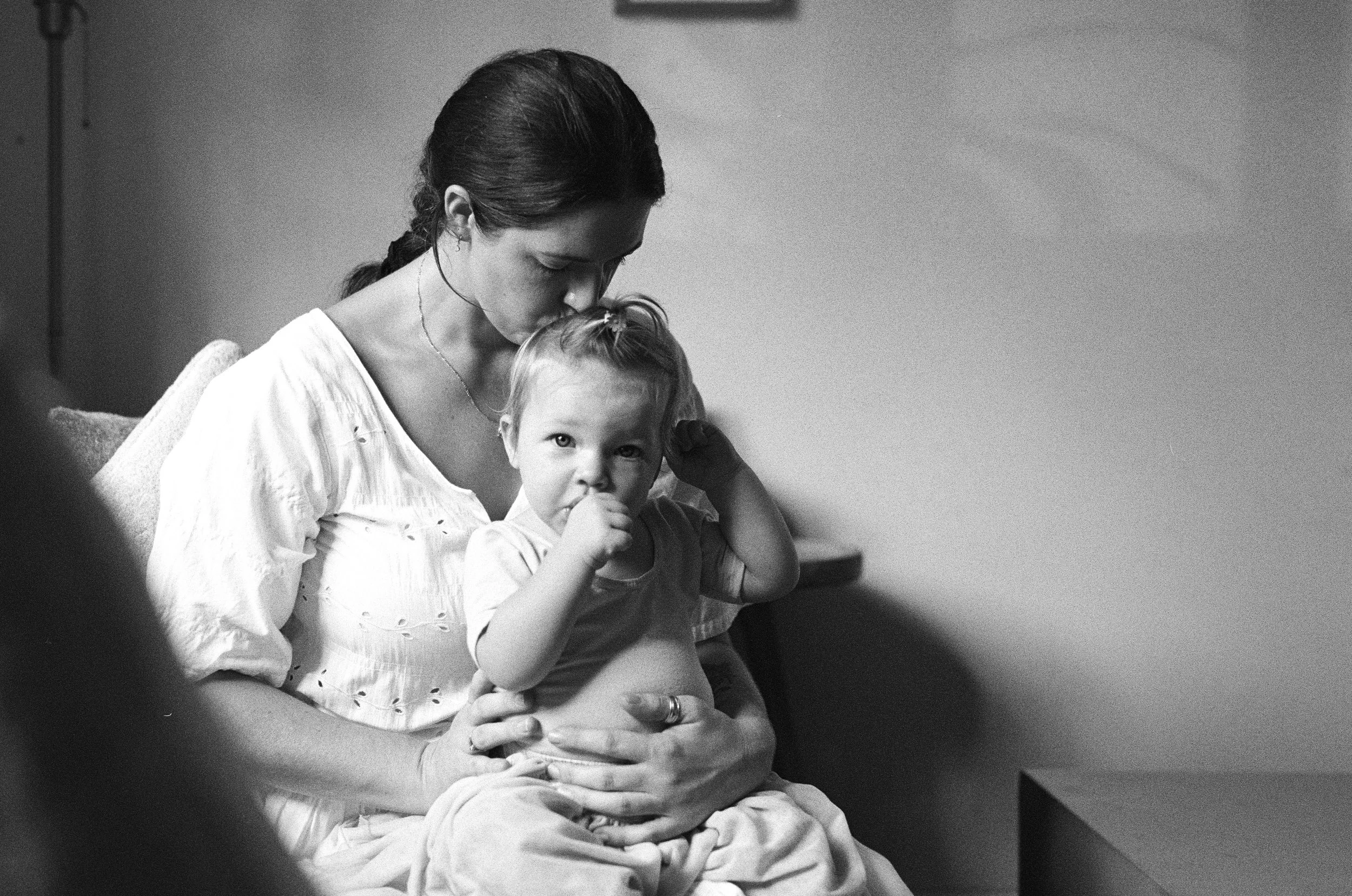 Black and white photo of a woman holding a young girl on her lap, with the woman kissing the girl's forehead, sitting on a couch in a room.