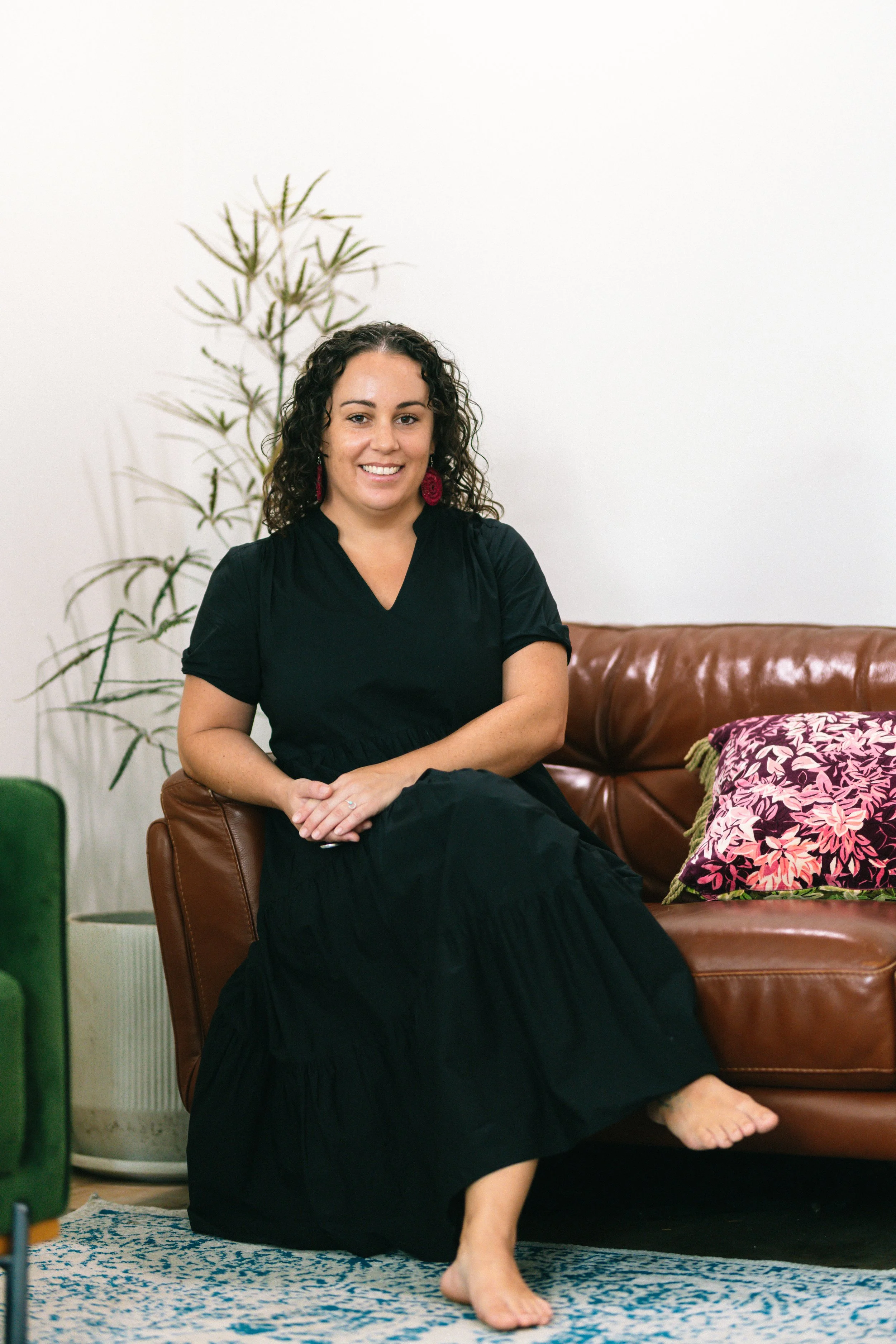 A woman with curly brown hair smiling, seated on a brown leather couch with a pink floral cushion, in a room with a white wall and a tall green plant in the background.