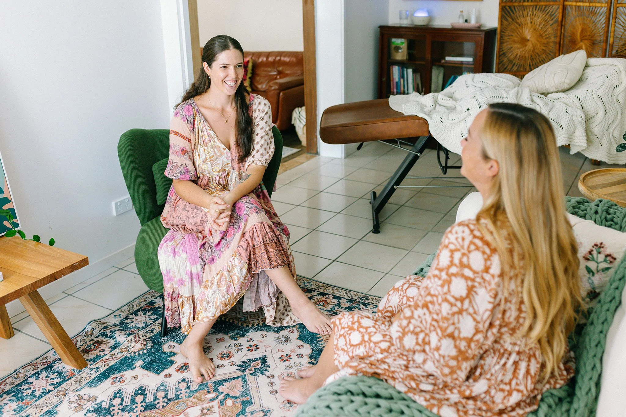Two women sitting and talking in a cozy, decorated living room with a bed and bookshelf visible in the background.