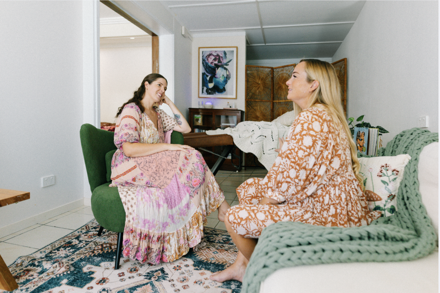 Two women having a conversation in a cozy living room, sitting on different chairs, with a colorful rug and wall art in the background