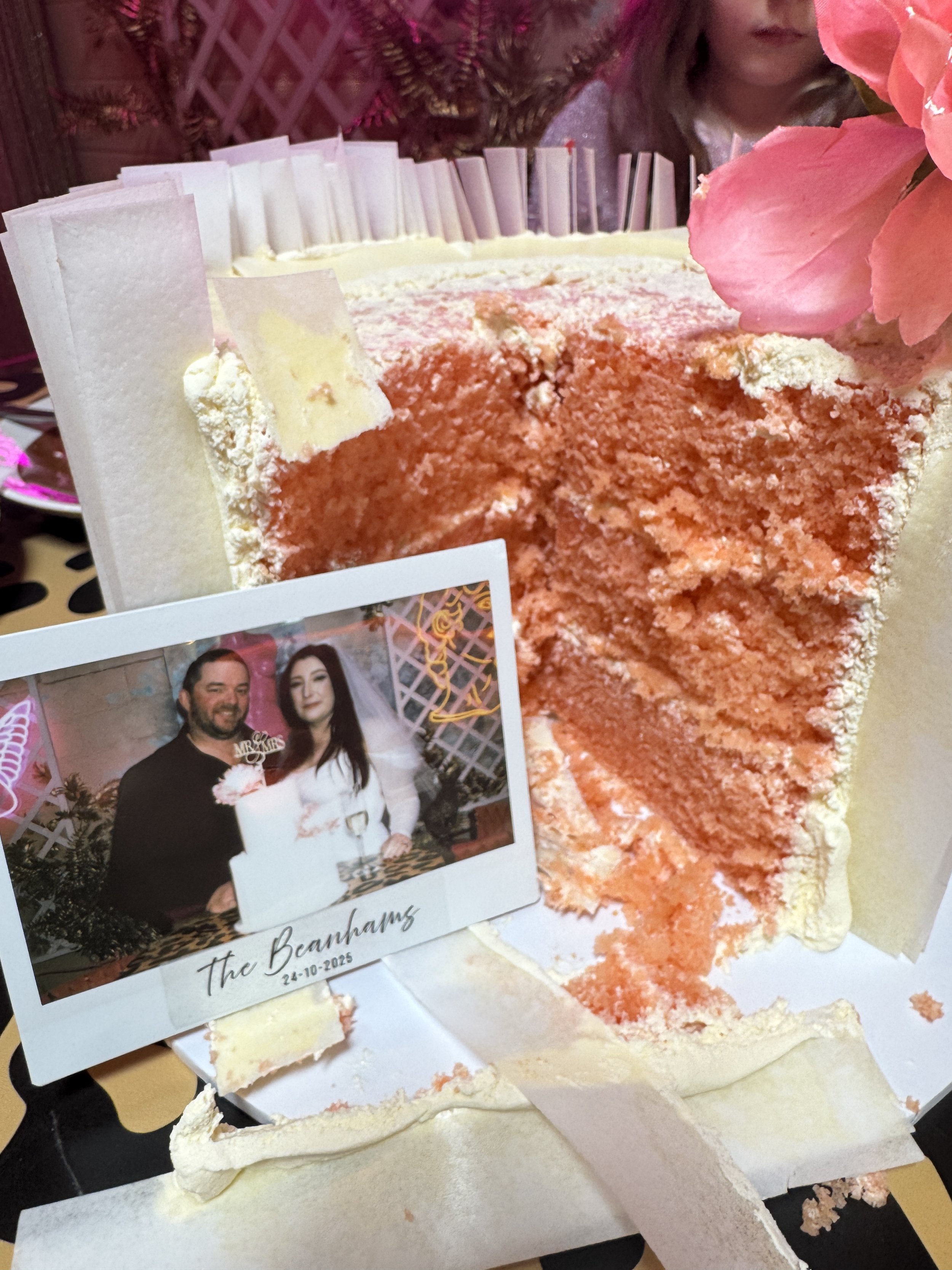 Slice of pink layered cake with pink flower decoration, partially cut, with a wedding photo of a couple on the table, and a background with pink neon lights and decorative elements.