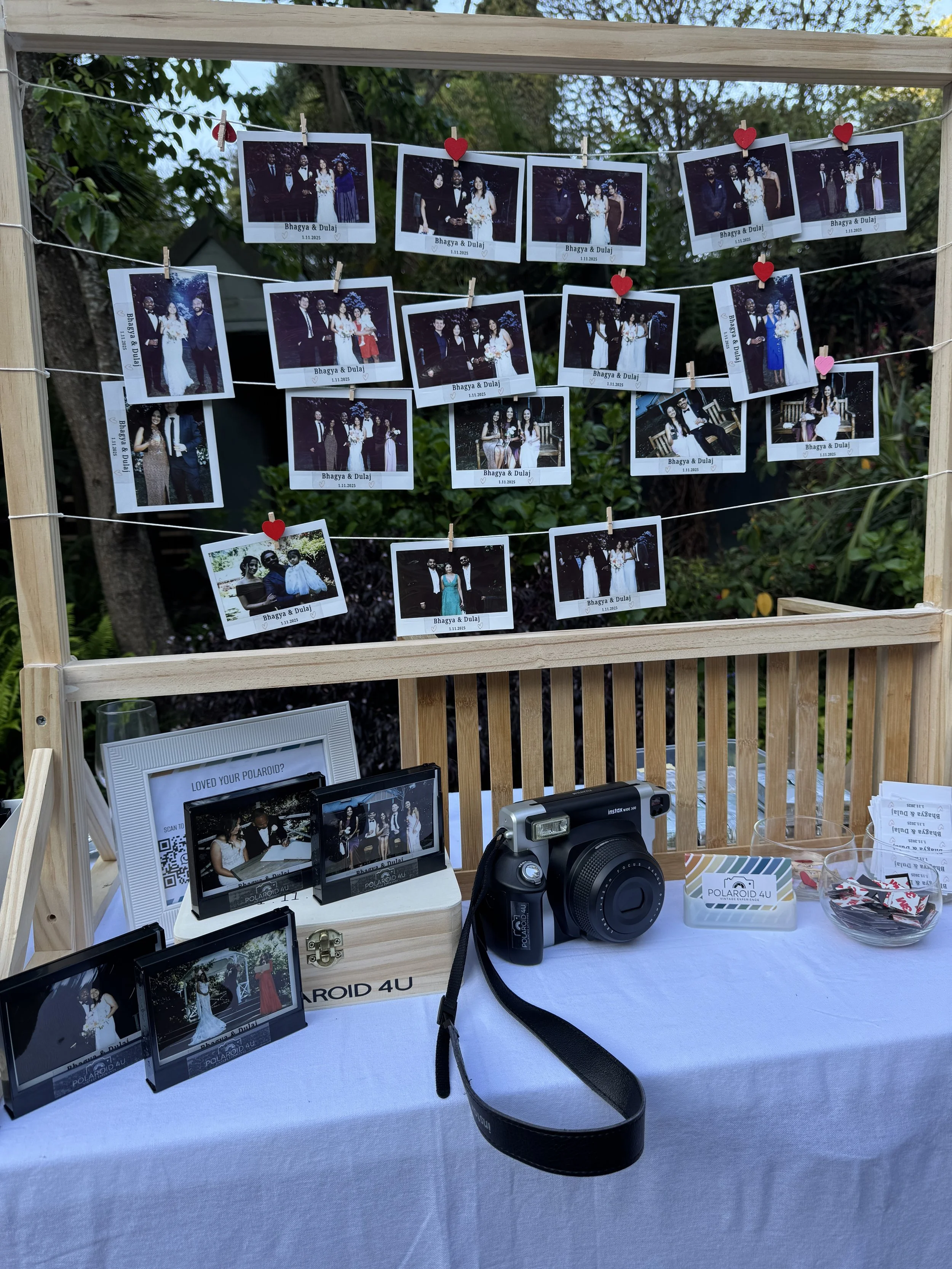 Photo of a wedding photo display table with group photos hanging on a frame, a camera, and picture displays on a table.