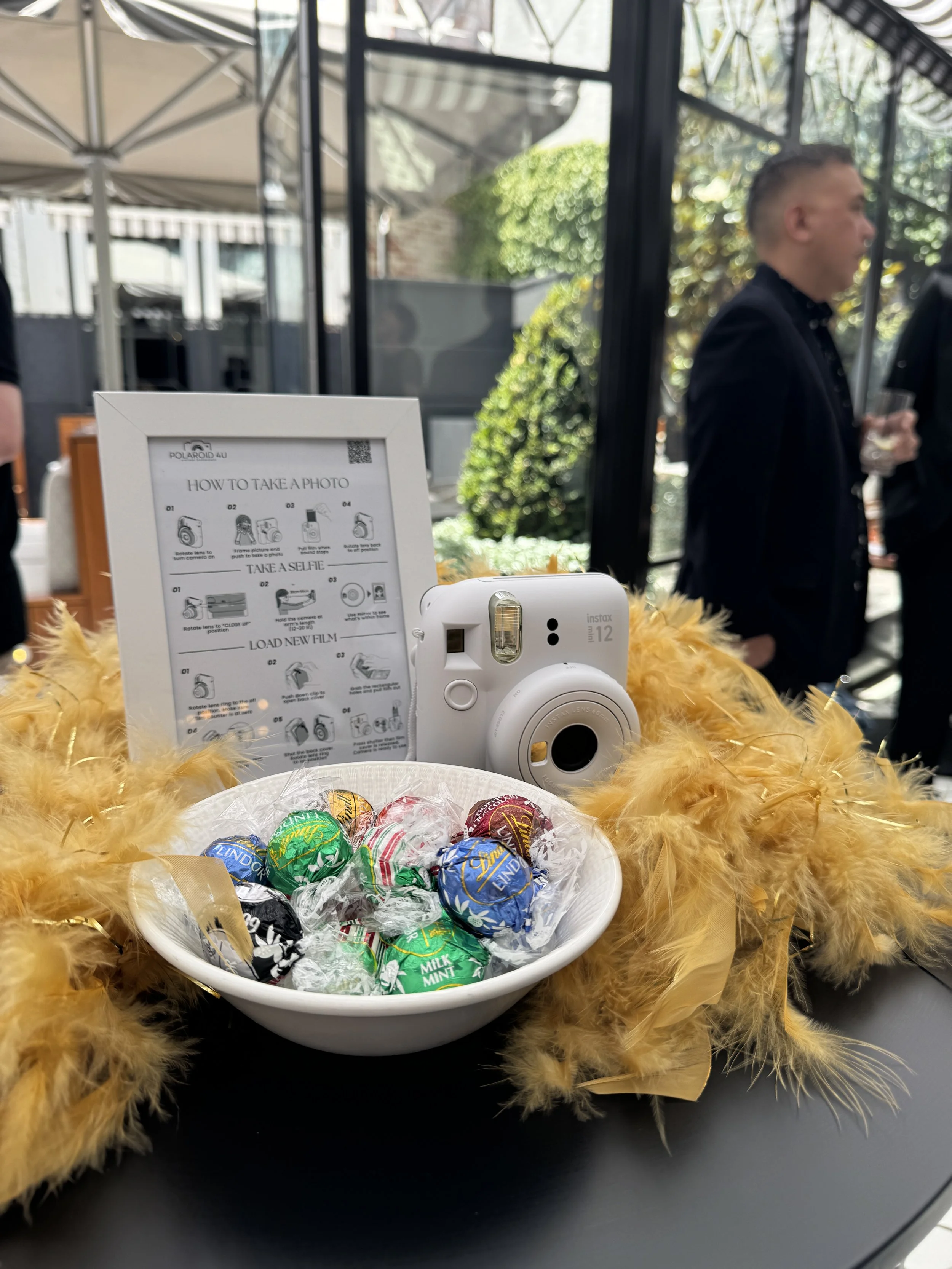 Polaroid camera, bowl of wrapped candies, and a sign providing instructions on how to take a photo at a photo booth, set on a table decorated with yellow feathers at an outdoor event.