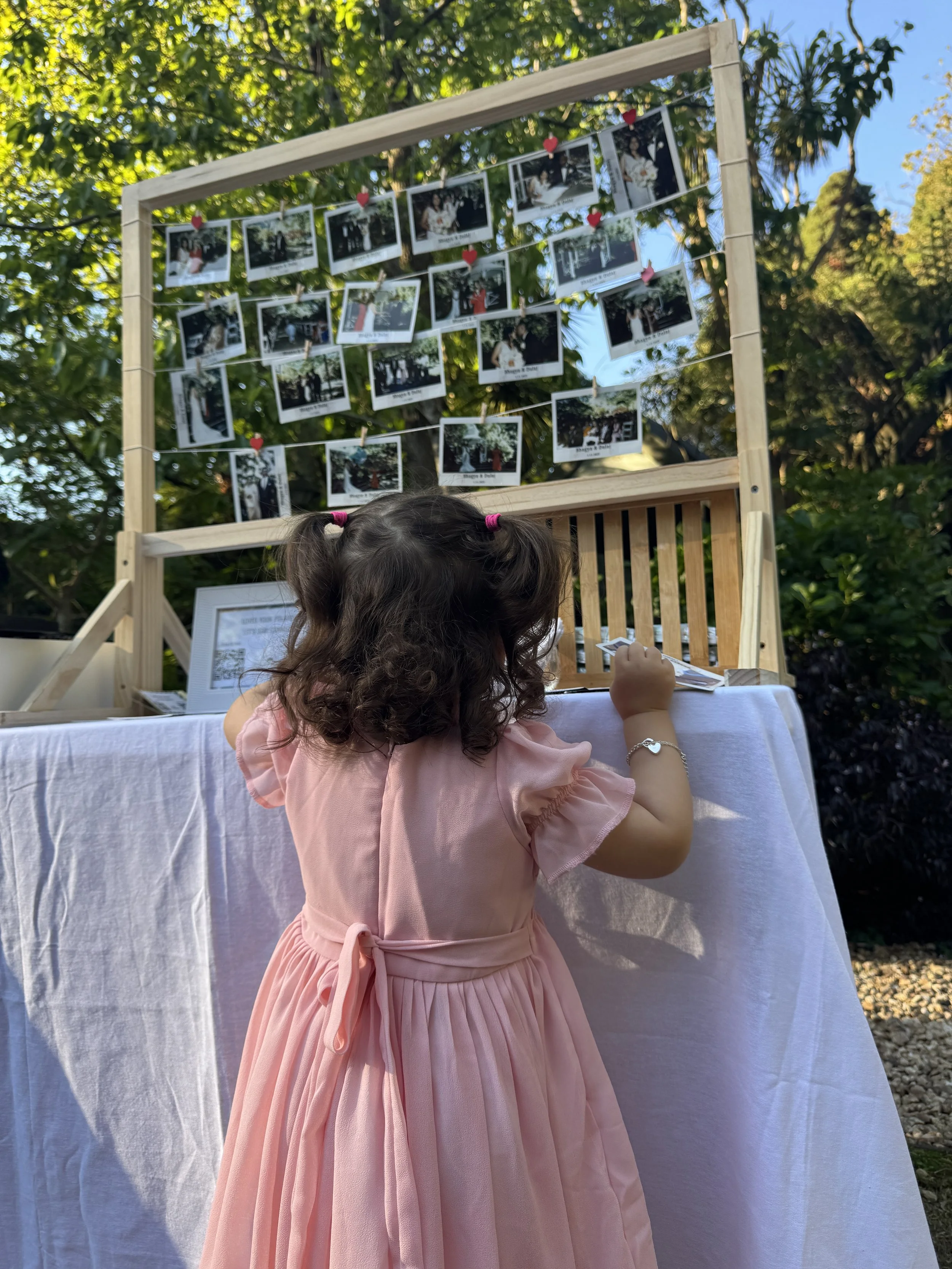 A young girl with curly brown hair in pigtails wearing a pink dress, standing at a table outdoors, writing or drawing on a piece of paper, with a display of photographs hanging on a wooden frame in the background.