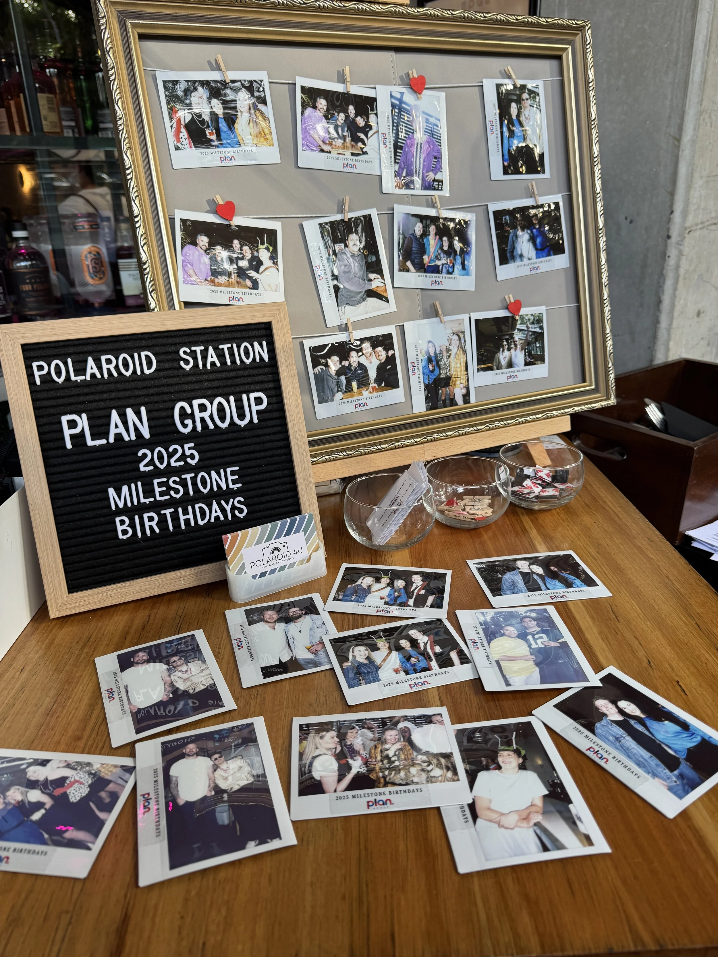 A wooden table with several printed photographs scattered on it, a black felt message board with white letters saying 'Polaroid Station Plan Group 2025 Milestone Birthdays', three glass bowls filled with small paper items, and a corkboard with a stri