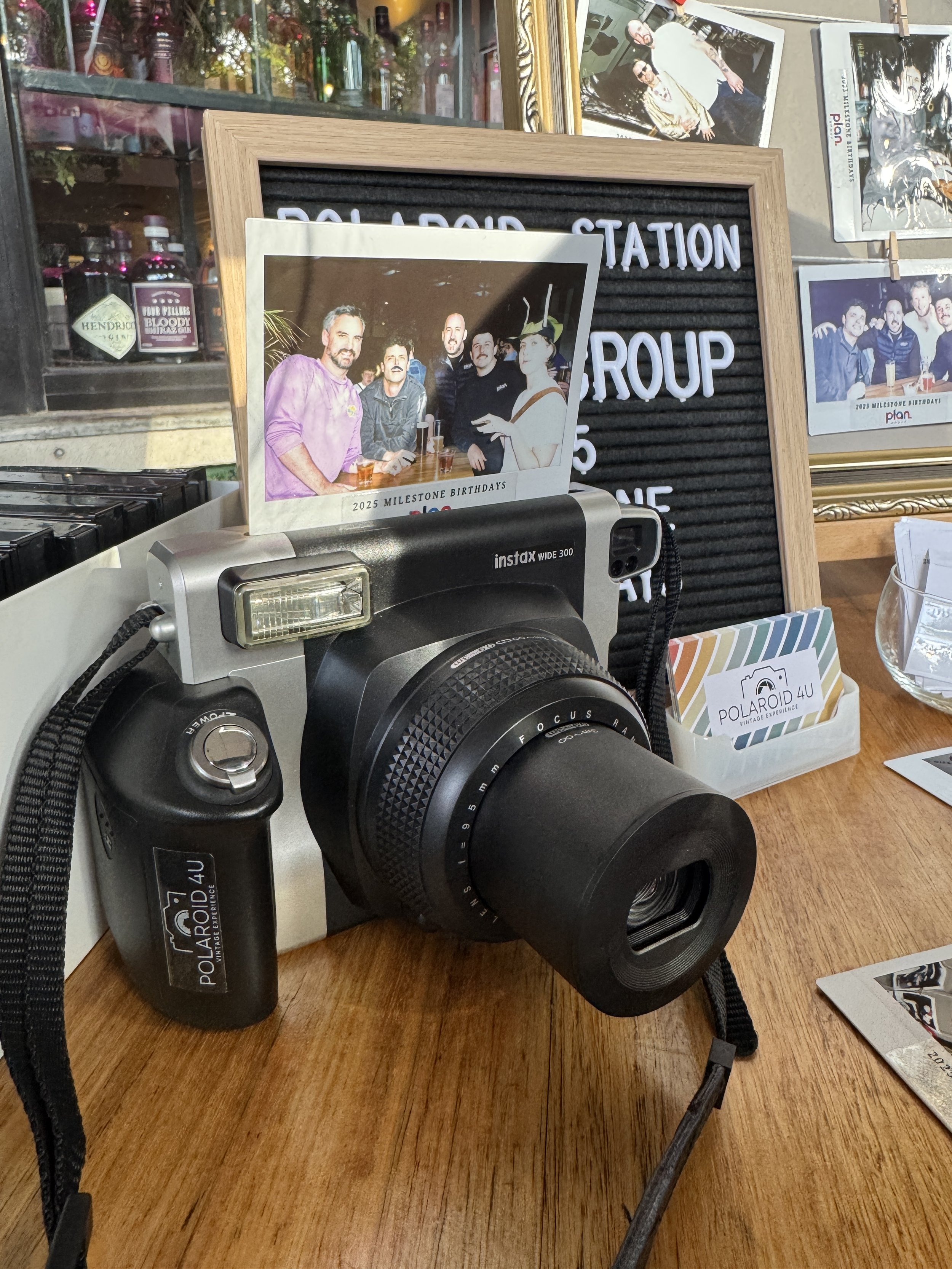 A vintage Polaroid camera on a wooden table with a photo of six people, various framed photographs, a sign that reads 'Pollard Station Group,' and a small box labeled 'Polaroid 4U.'