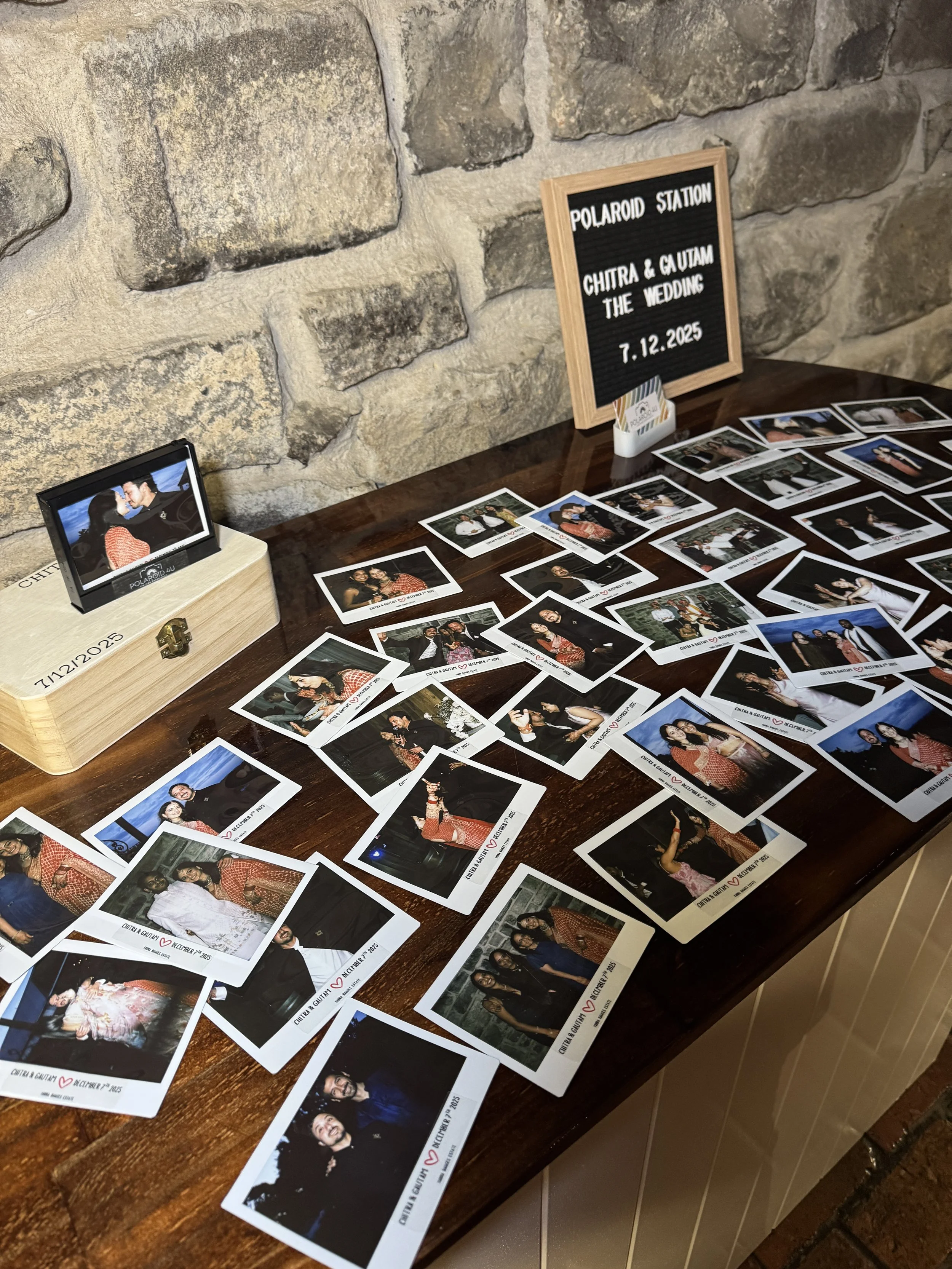 A table displays numerous Polaroid photos of people at a wedding, with a framed sign reading 'Polaroid Station, Chitra & Gautam, The Wedding, 7.12.2025' against a stone wall background.