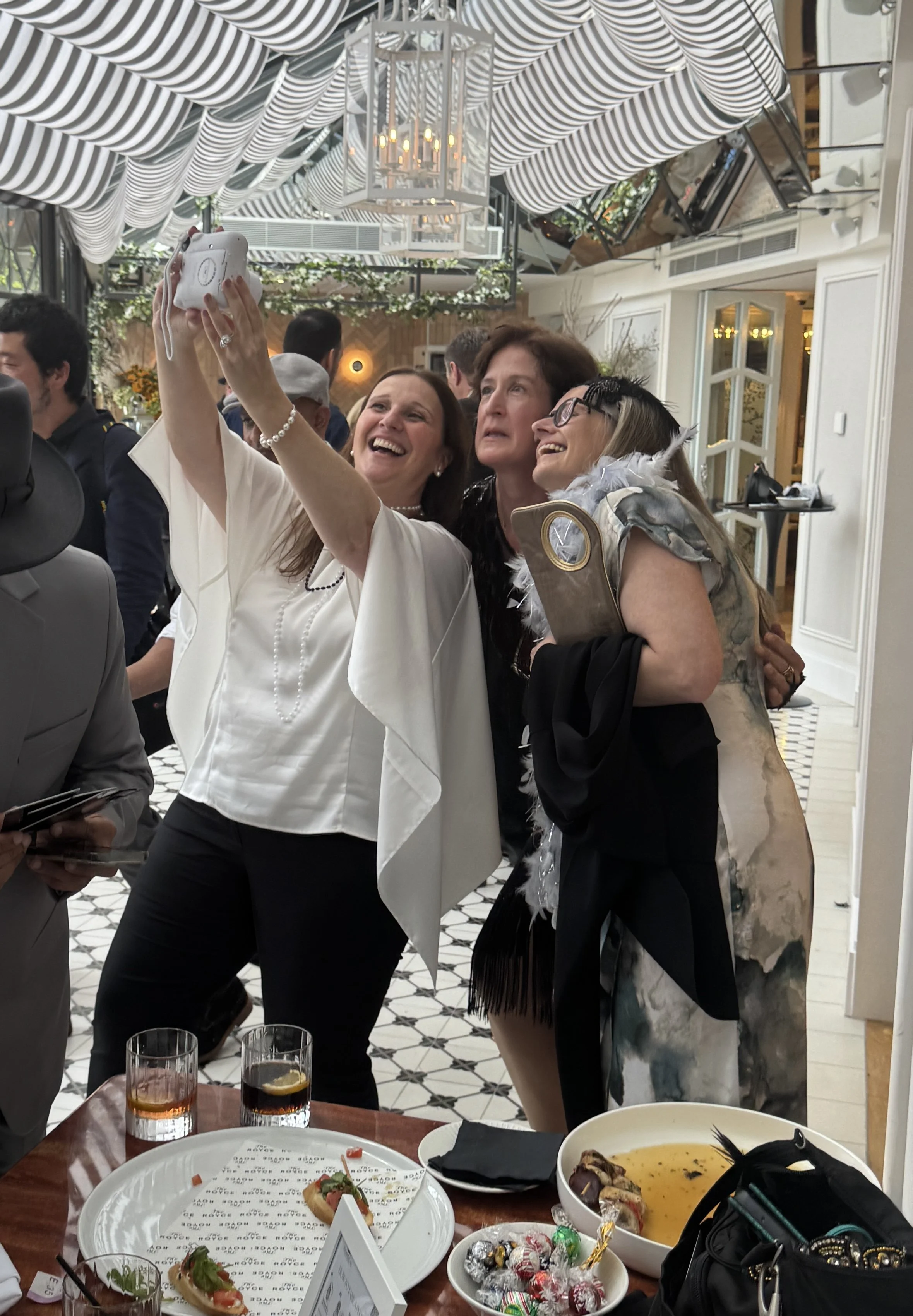 Three women taking a selfie at a social gathering or celebration, with a decorated ceiling and food on the table in the foreground.
