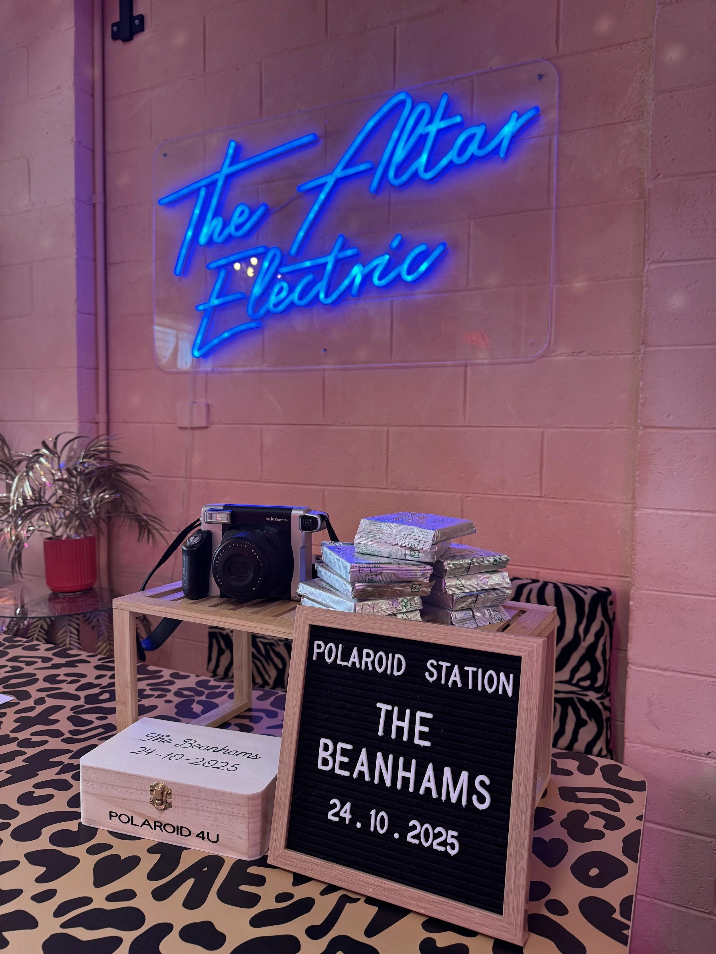 Decorative setup at a Polaroid station with a vintage camera, stacks of cash, a wooden box, a neon sign reading 'The Attar Electric,' and a message board displaying 'The Beanhams 24.10.2025' on a pink wall with leopard print tablecloth.