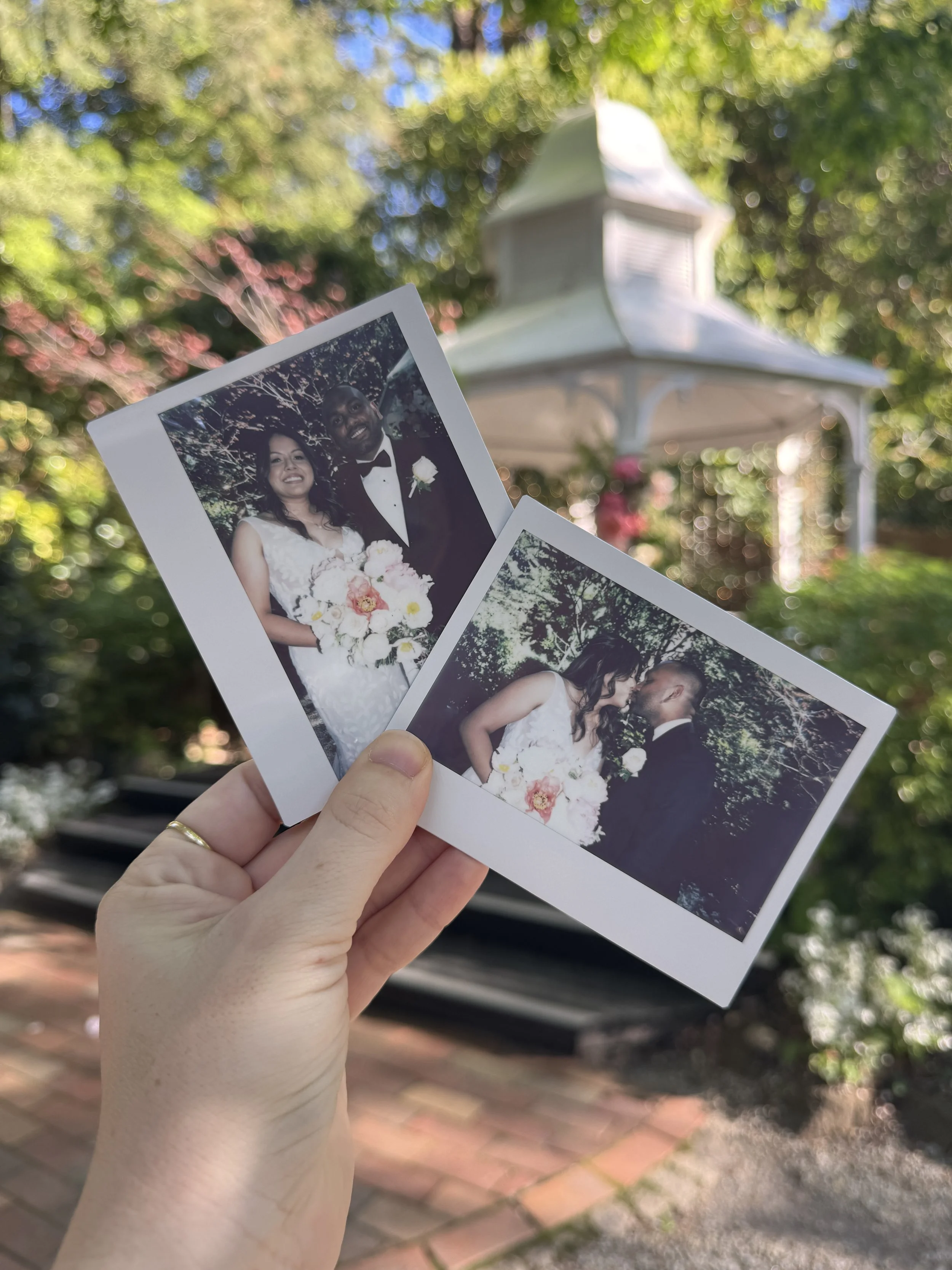 Hand holding two wedding photos outdoors with a white gazebo and green trees in the background.