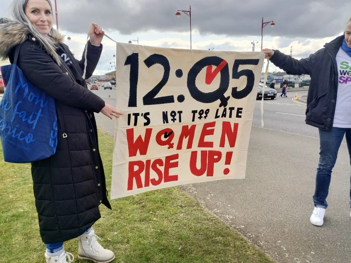 Two people holding a protest banner that reads "12:05 It's Not Too Late Women Rise Up!" outdoors near a road.
