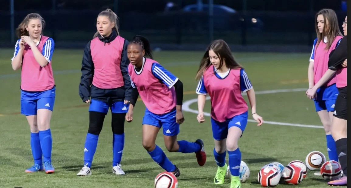 Group of young female soccer players in pink bibs and blue uniforms training on a field with soccer balls.