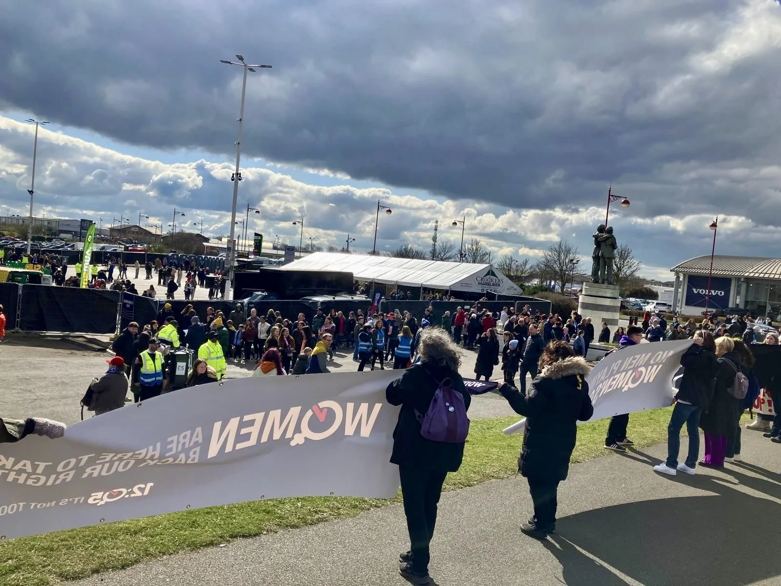 A group of people gathered outdoors holding a large banner that reads 'WOMEN' in large letters. The event appears to be a protest or rally, with additional crowd members visible in the background. There are tents and a statue visible, along with clou