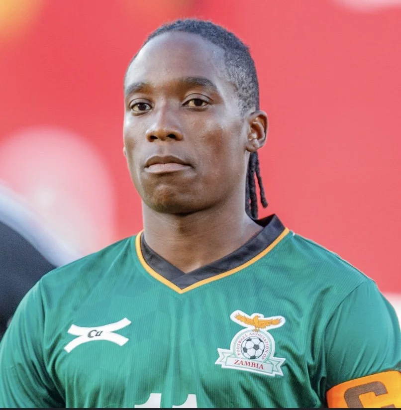 Soccer player wearing a green Zambia national team jersey with a captain's armband, standing against a red background.
