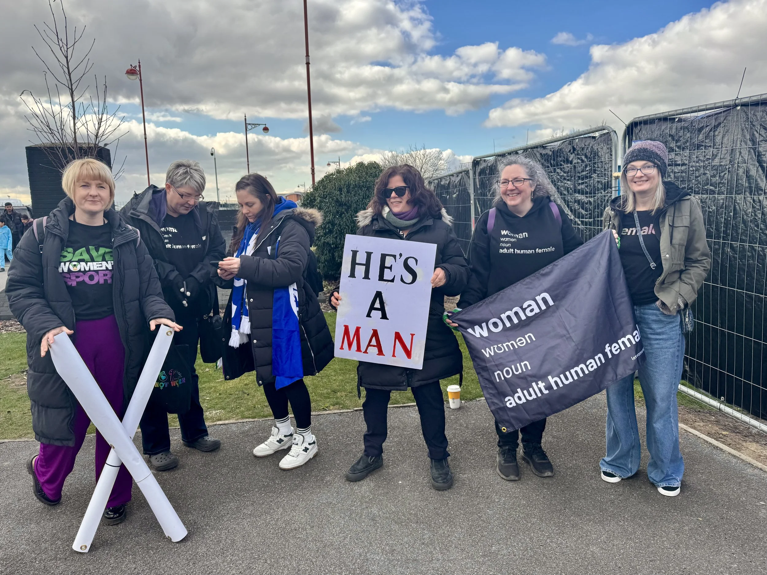 Group of people, some holding signs and banners, in an outdoor setting with partially cloudy skies.
