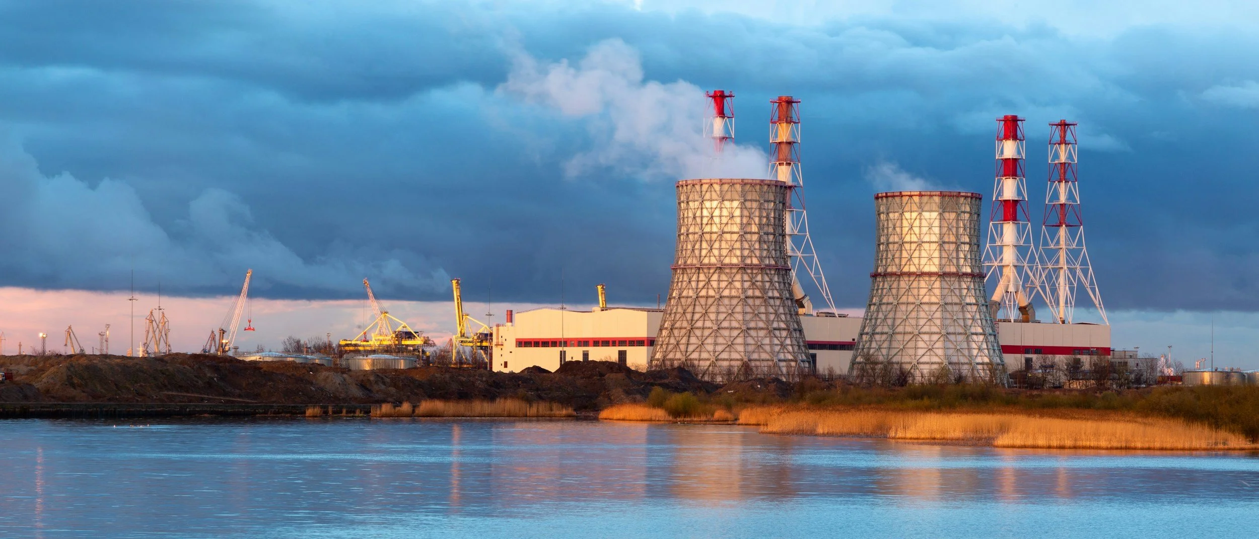 A nuclear power plant situated near a body of water with cooling towers emitting steam under a cloudy sky during sunset.