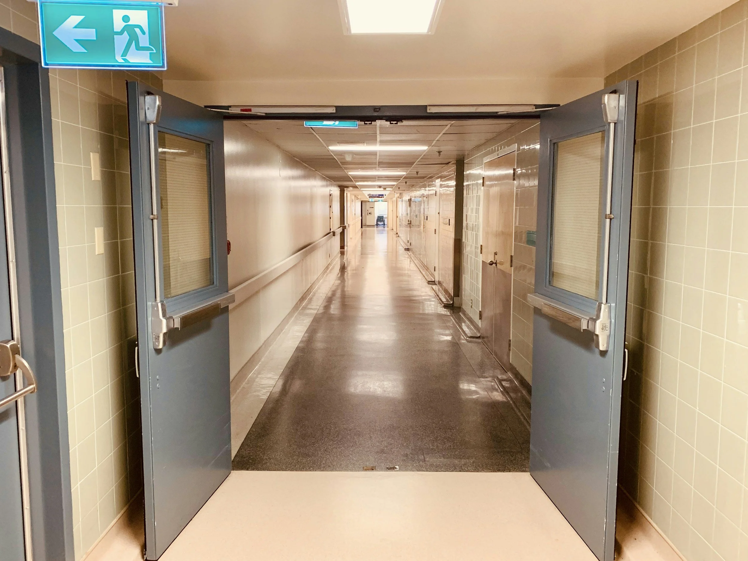 Long hospital corridor with closed doors on each side and emergency exit sign above the entrance.