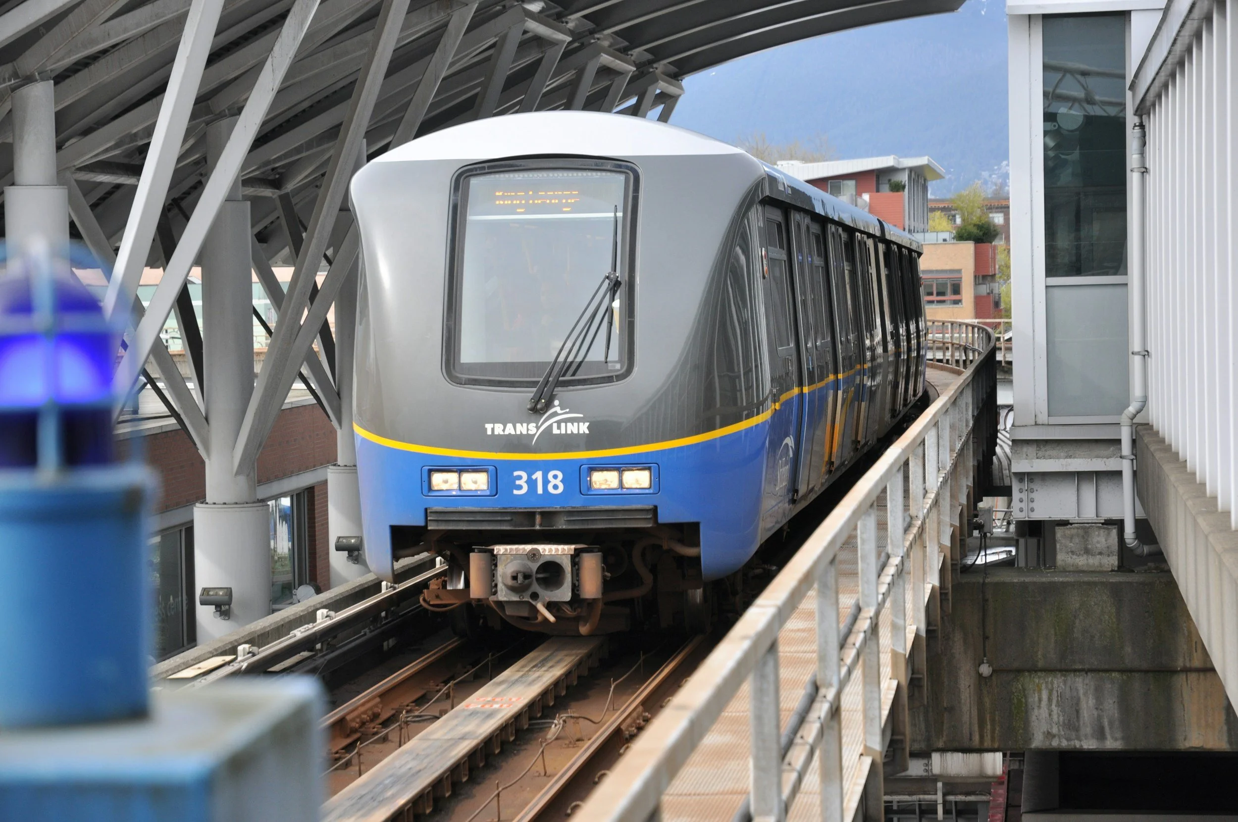 A blue and gray TransLink SkyTrain traveling on an elevated track at a station with modern architecture and a mountainous background
