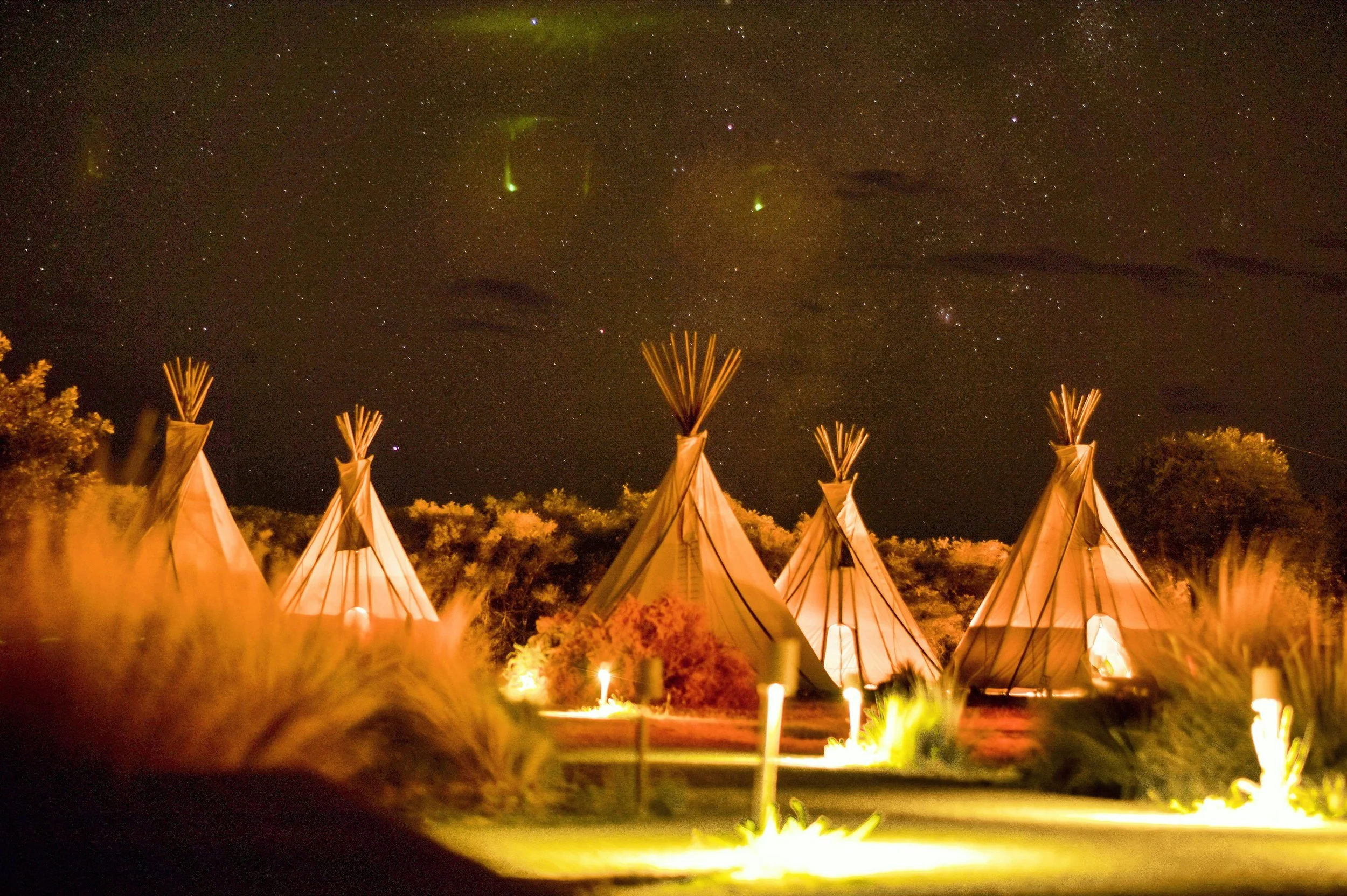 Nighttime scene with five Tipi tents illuminated from within, set against a starry sky with visible the Milky Way and green auroras, surrounded by bushes and trees.