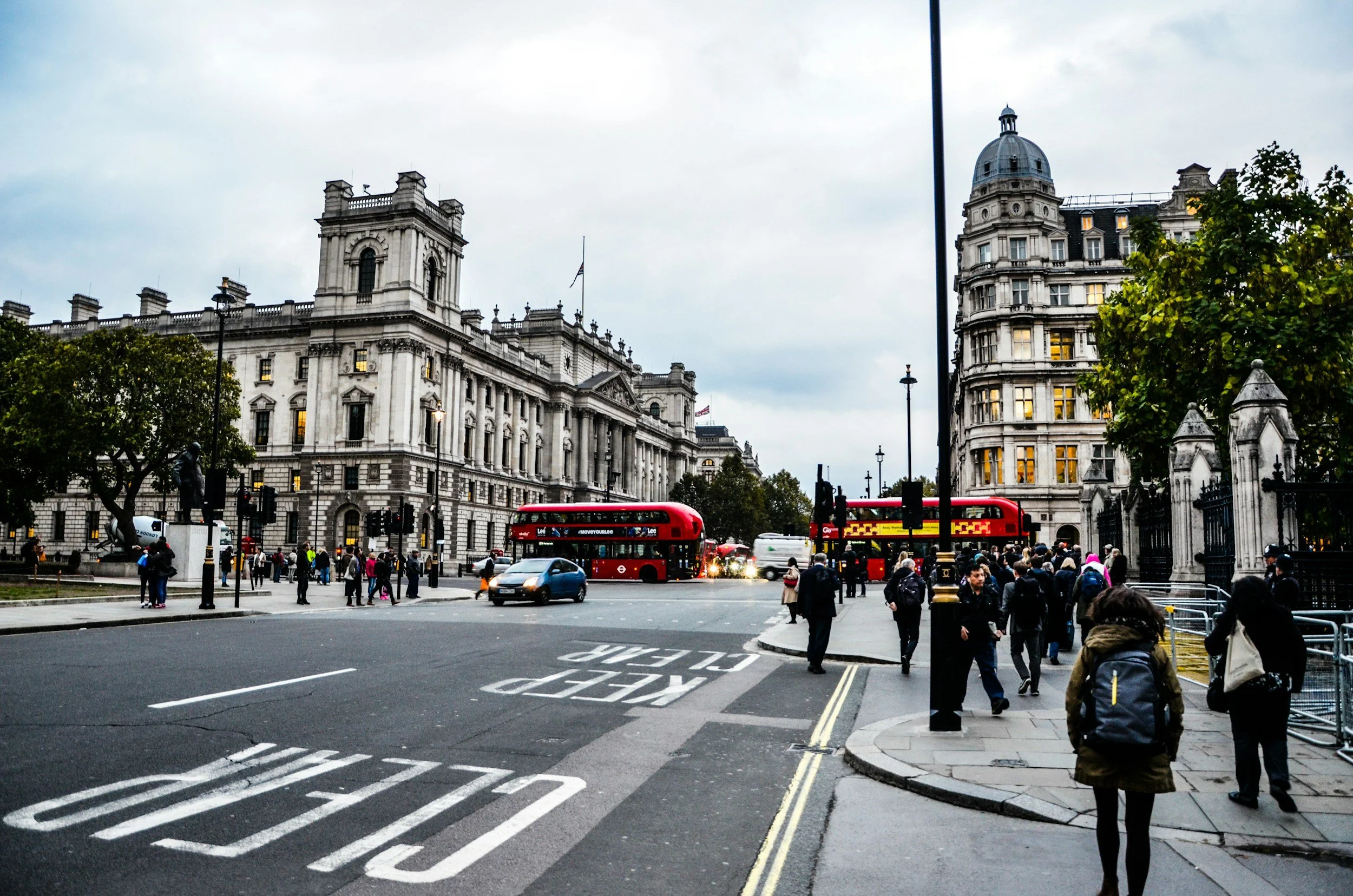Street scene in London with historic buildings, pedestrians, and red double-decker buses.