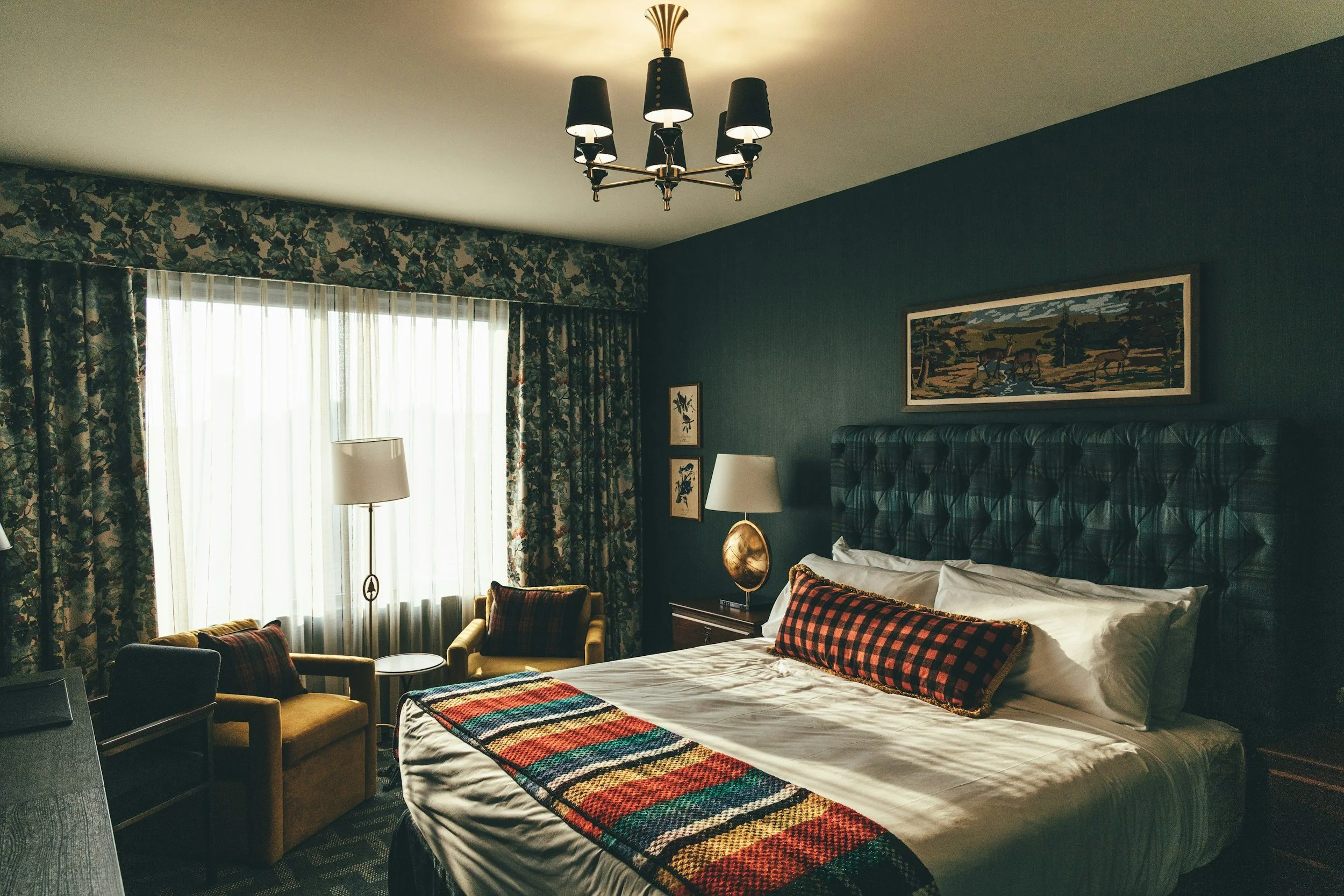 A hotel room with a large bed, a plaid headboard, and a colorful striped blanket. There are two armchairs with plaid pillows near a window with floral curtains. The room has mixed artwork and a chandelier hanging from the ceiling.
