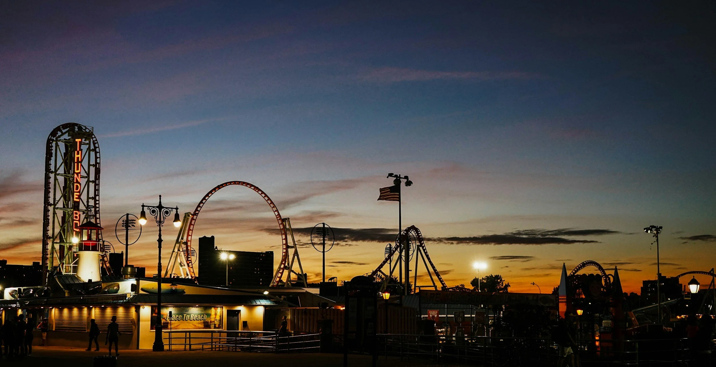 Amusement park with roller coasters and rides during sunset, silhouetted against the colorful sky, with some people walking and booths in the foreground.