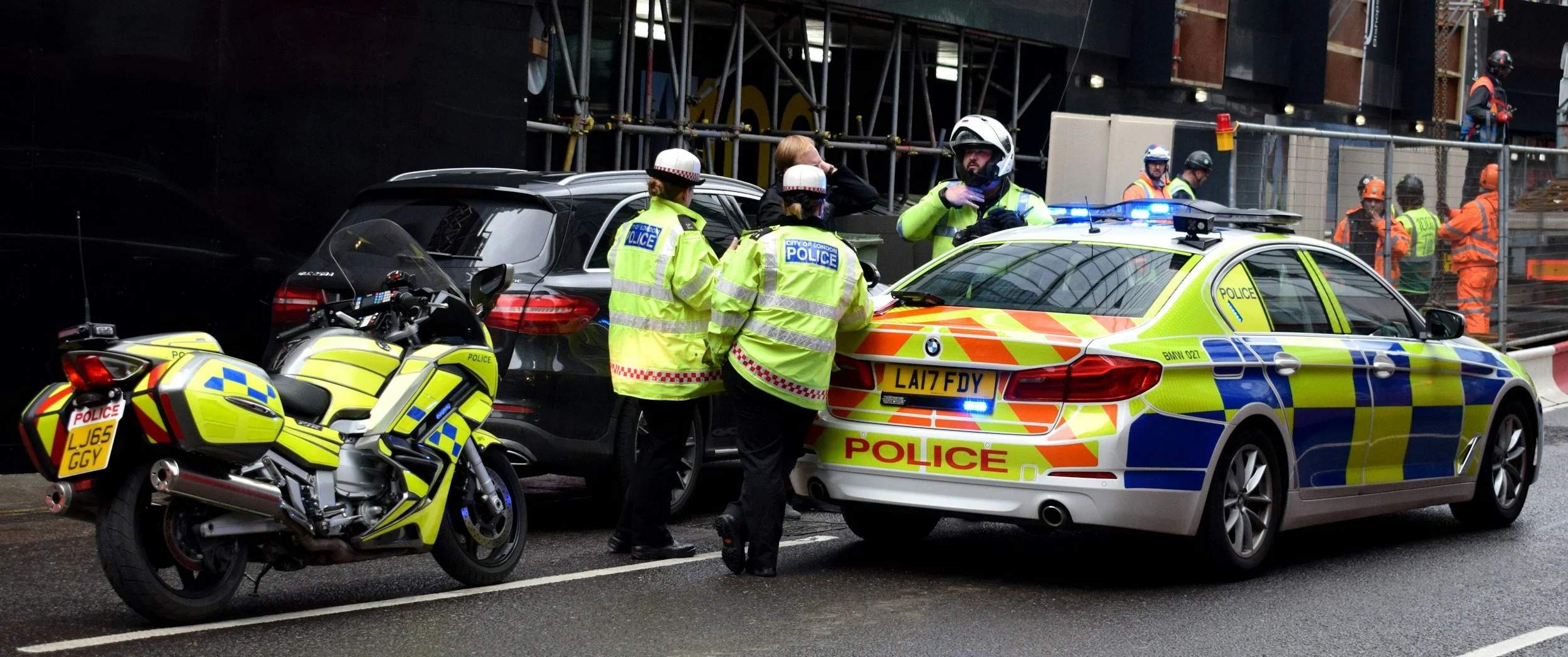 Police officers in high-visibility jackets and helmets stand near police cars and a motorcycle on a city street, with construction workers and scaffolding in the background.