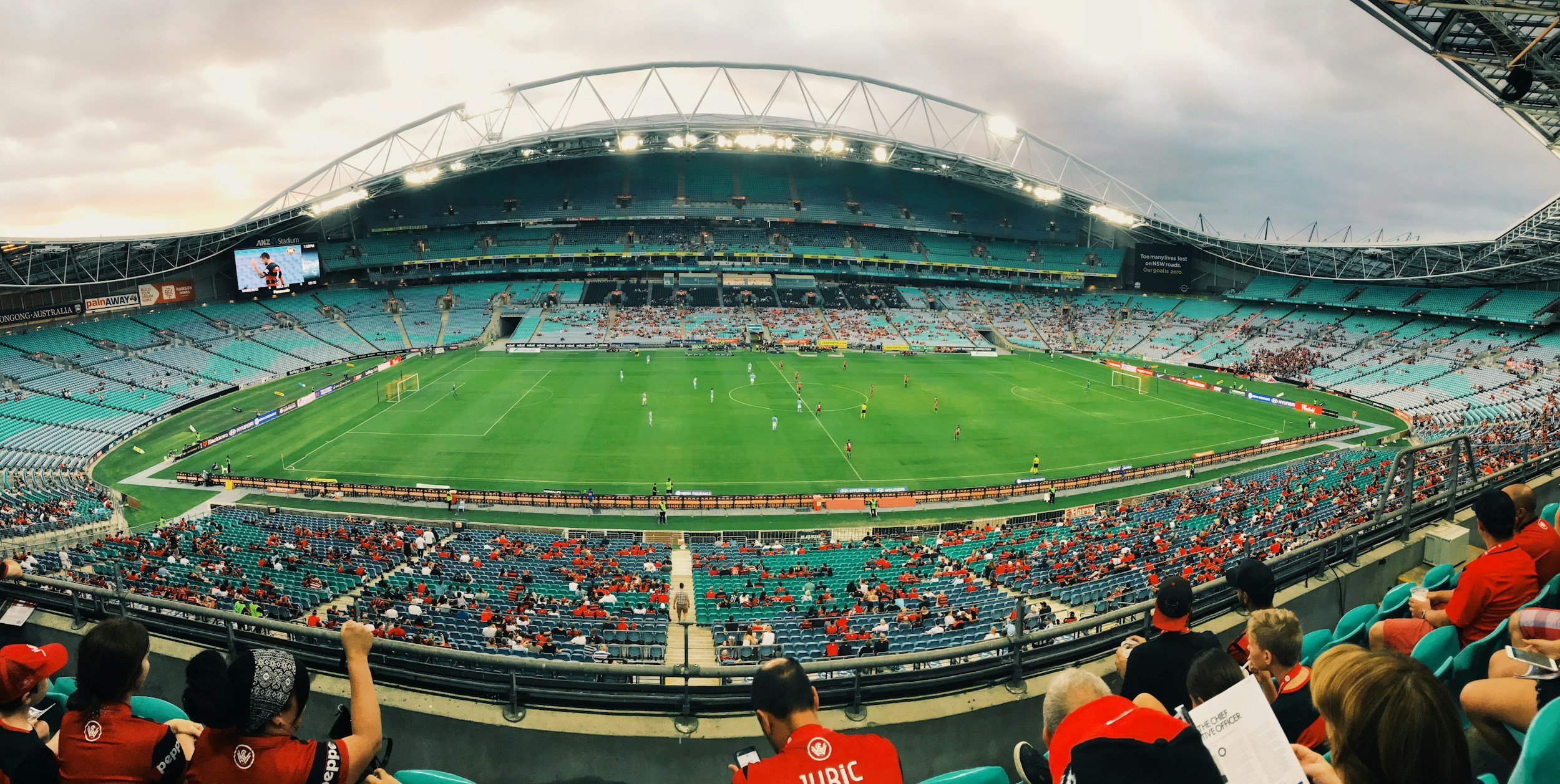 The image shows a nearly empty stadium with a soccer match in progress, featuring players on the field, spectators in the stands, and overcast skies overhead.