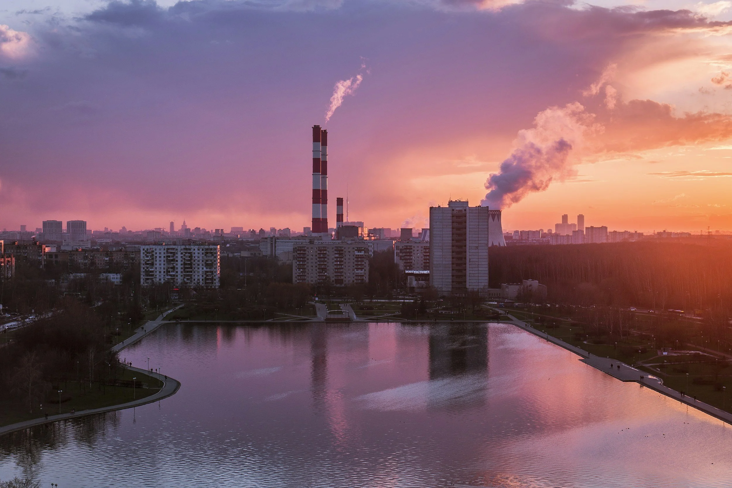 Sunset view of a city with industrial smokestacks emitting smoke, a body of water in the foreground, and high-rise buildings in the background.