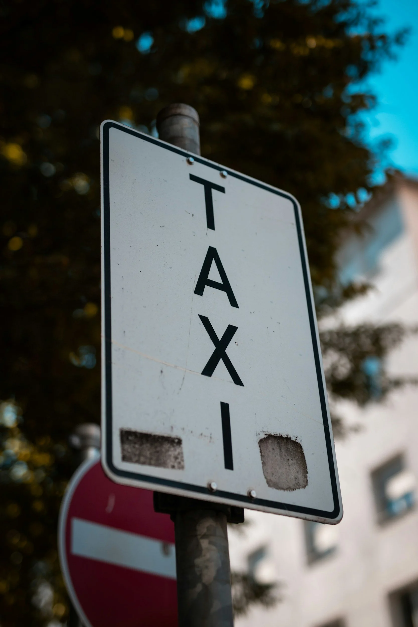 A weathered parking sign reading 'TAXI' mounted on a pole, with a blurred building and trees in the background.